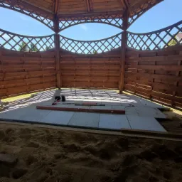 Montaż szarej kostki brukowej w drewnianej altanie, poziomice i gumowy młotek na ułożonej powierzchni, podsypka piaskowa widoczna na pierwszym planie.