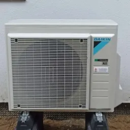 Zewnętrzna jednostka pompy ciepła Daikin, model R-32, zamontowana na betonowych podstawach przy jasnej ścianie budynku.