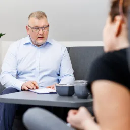 Mężczyzna w okularach siedzi przy stoliku z notatnikiem i długopisem, rozmawiając z osobą siedzącą naprzeciwko, widoczną od tyłu, w tle roślina doniczkowa.