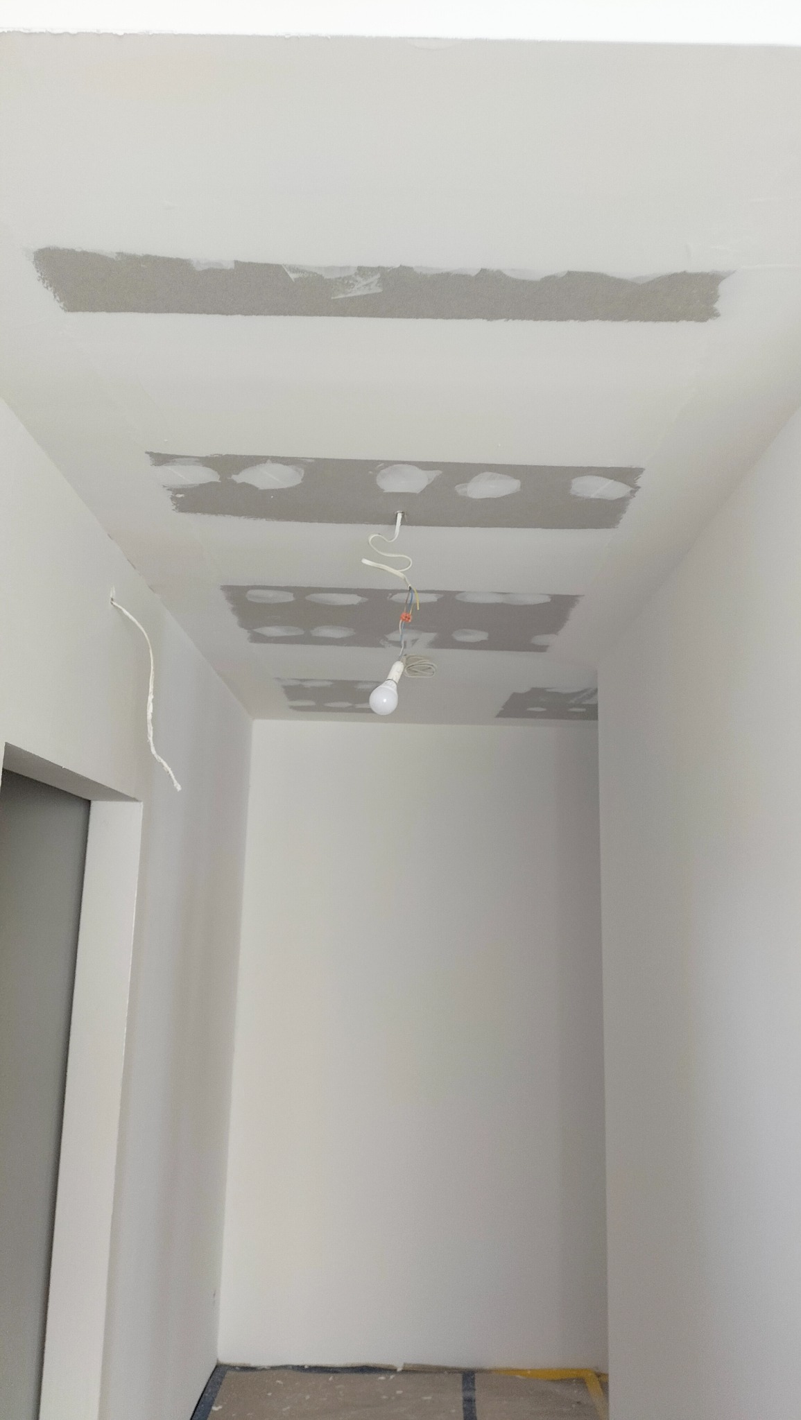 Wnętrze w trakcie prac wykończeniowych: sufit z widocznymi fragmentami szpachlowanej płyty, wisząca żarówka na kablu, białe ściany w trakcie malowania.