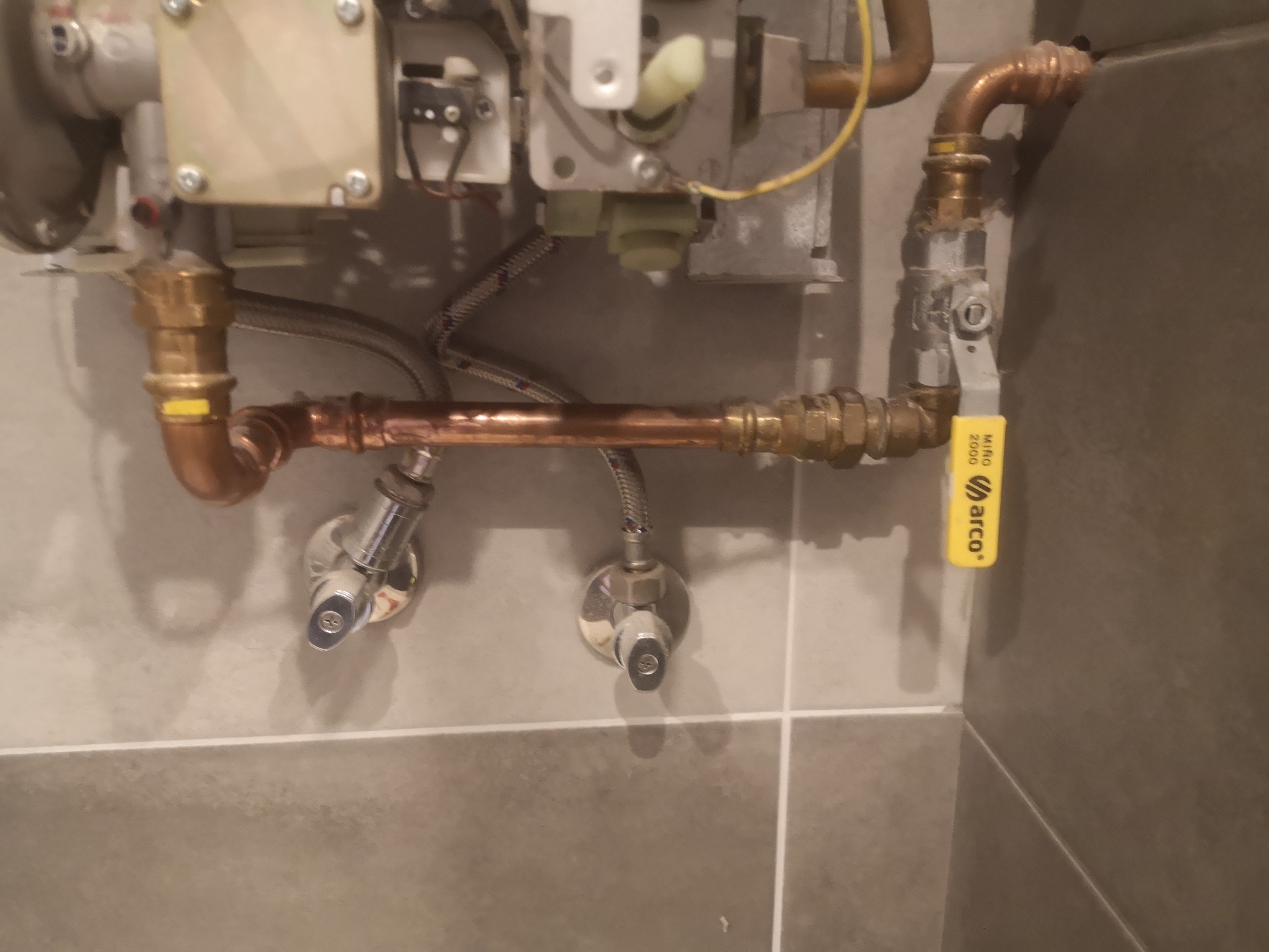 Fragment instalacji gazowej z miedzianymi rurami, zaworami i żółtym oznaczeniem Arco 2000, zamontowanej na szarej, kafelkowanej ścianie.