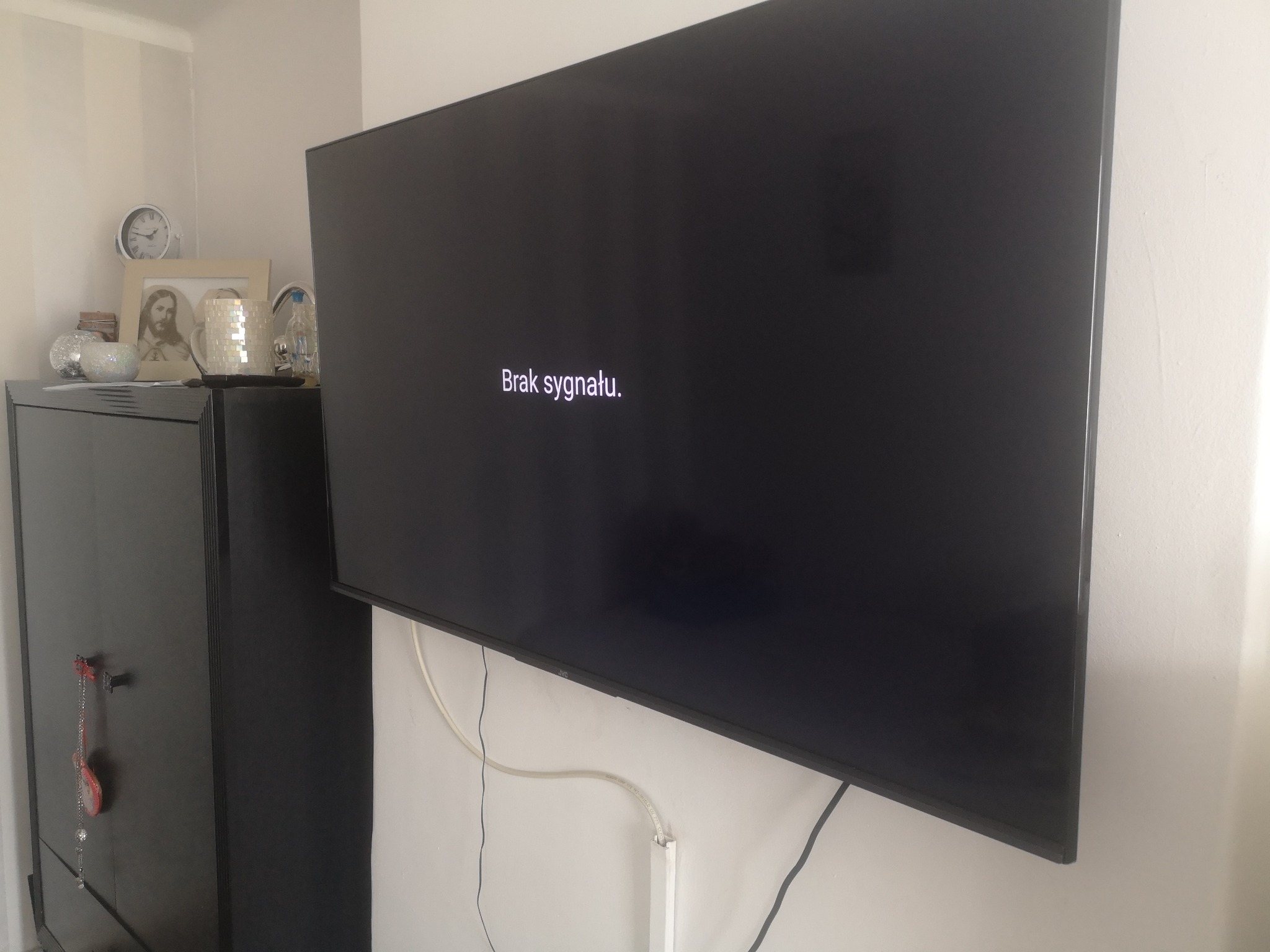 Telewizor LCD zawieszony na ścianie, wyświetlający komunikat 'Brak sygnału', z widocznymi kablami i szafką w tle.