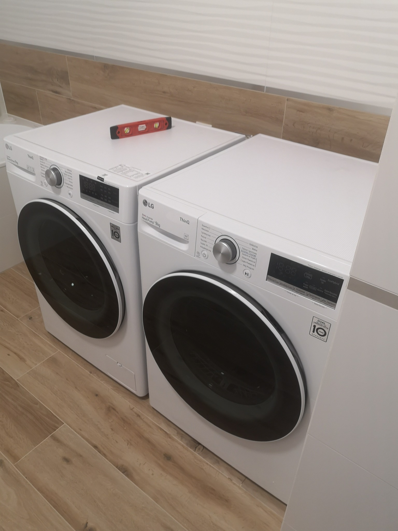 Dwie białe pralki LG ustawione obok siebie w łazience, poziomica spoczywająca na jednej z nich, wskazująca na prawidłowe wypoziomowanie, płytki imitujące drewno na podłodze i ścianie.