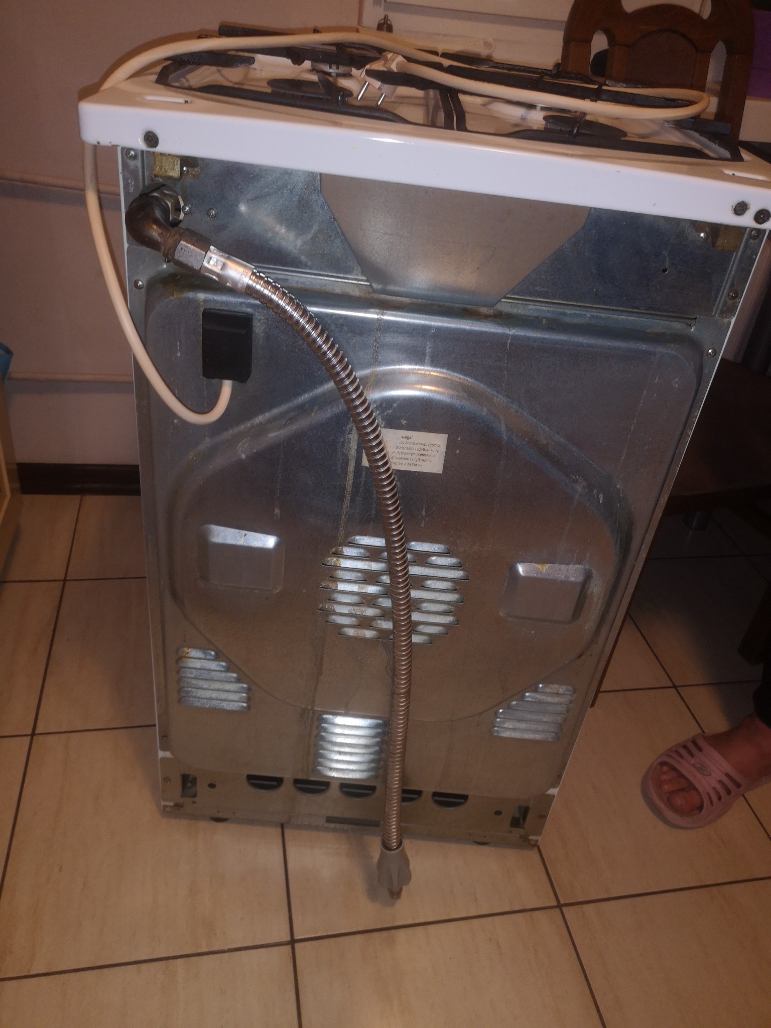 Tył kuchenki gazowej z podłączonym przewodem gazowym w metalowym oplocie, widoczne przyłącze i etykieta informacyjna na obudowie, na podłodze płytki i fragment stopy w klapku.