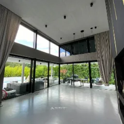 Jasnoszara podłoga w przestronnym, nowoczesnym salonie z panoramicznymi oknami wychodzącymi na ogród z basenem; widoczne minimalistyczne oświetlenie sufitowe i zasłony w kolorze beżowym.