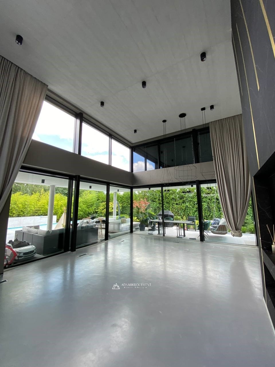 Jasnoszara podłoga w przestronnym, nowoczesnym salonie z panoramicznymi oknami wychodzącymi na ogród z basenem; widoczne minimalistyczne oświetlenie sufitowe i zasłony w kolorze beżowym.