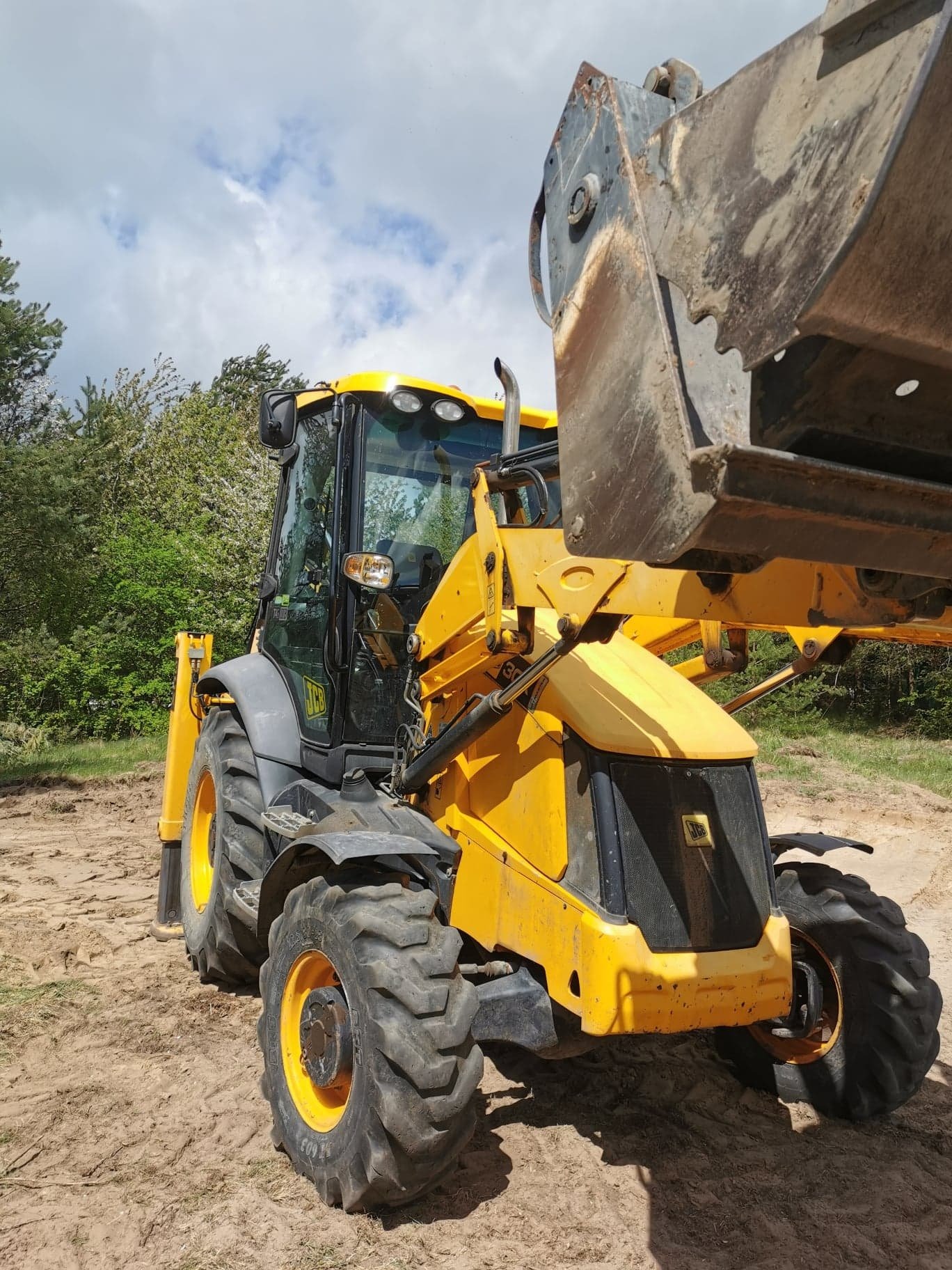 Żółto-szara koparko-ładowarka JCB na piaszczystym terenie, z podniesioną łyżką i widocznym lasem w tle pod pochmurnym niebem.