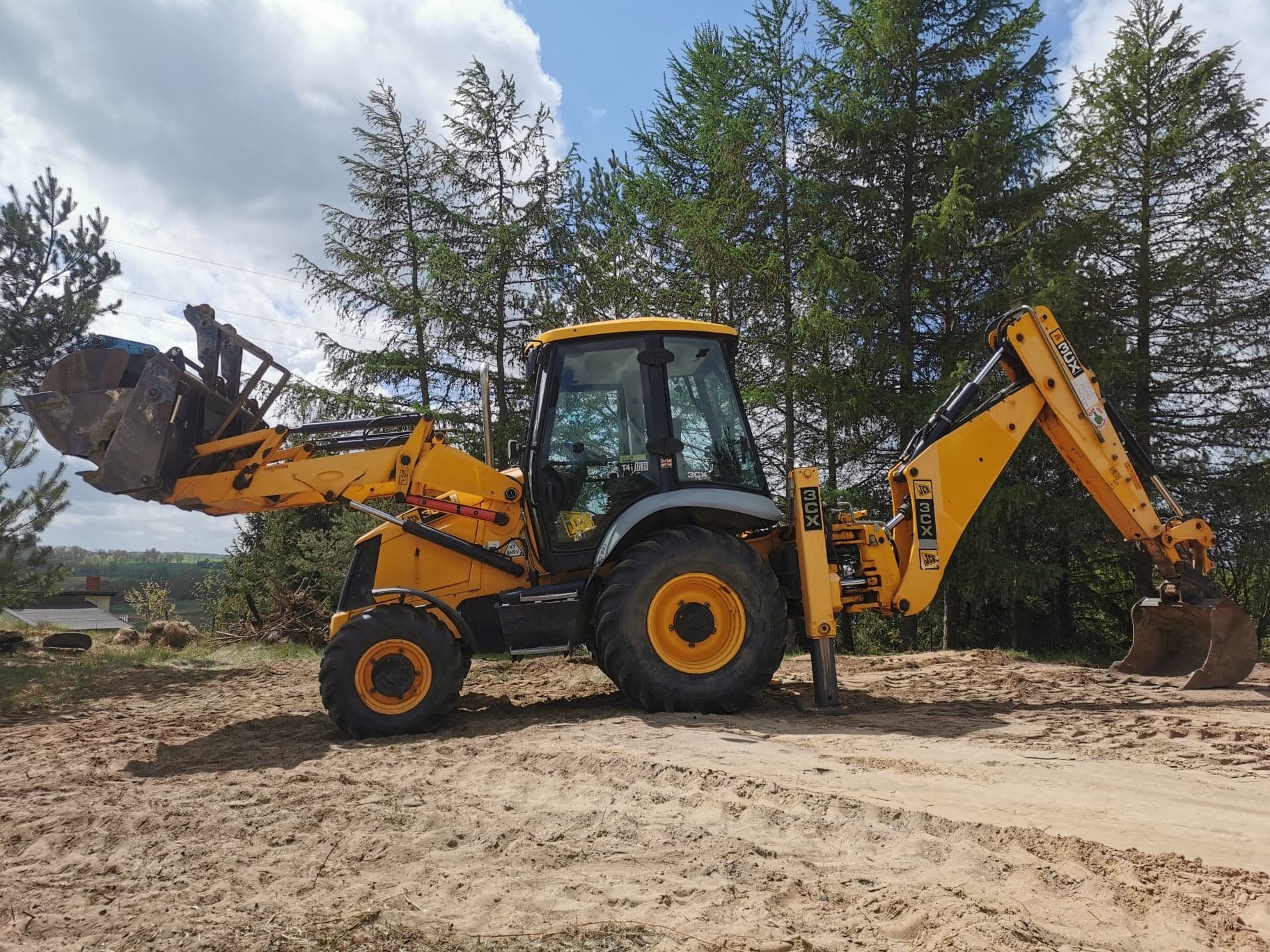 Żółto-szara koparko-ładowarka JCB 3CX na piaszczystym terenie, z łyżką podniesioną do góry i wysuniętym ramieniem koparki, w tle drzewa iglaste i pochmurne niebo.