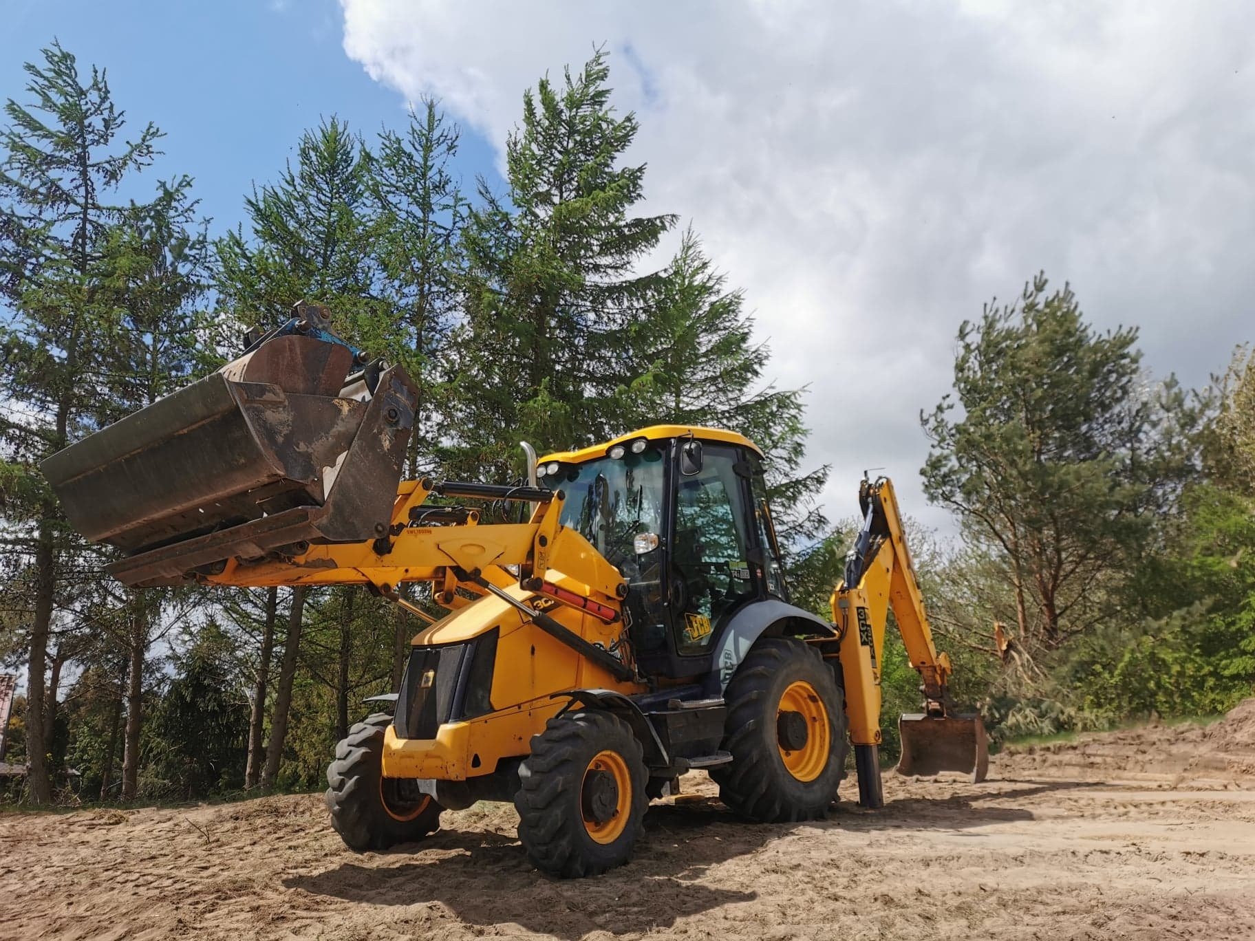 Żółta koparko-ładowarka JCB na piaszczystym terenie, z podniesioną łyżką i widocznymi śladami opon, na tle drzew i pochmurnego nieba.
