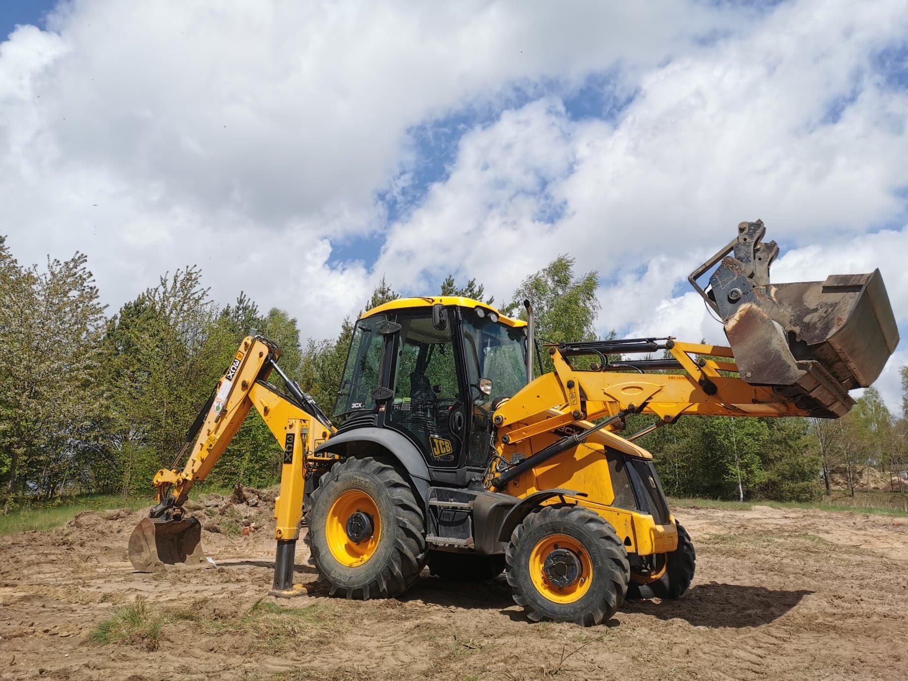 Żółto-szara koparko-ładowarka JCB na placu budowy, z podniesioną łyżką i wysuniętym ramieniem kopiącym, na tle ziemi i drzew pod błękitnym niebem z chmurami.