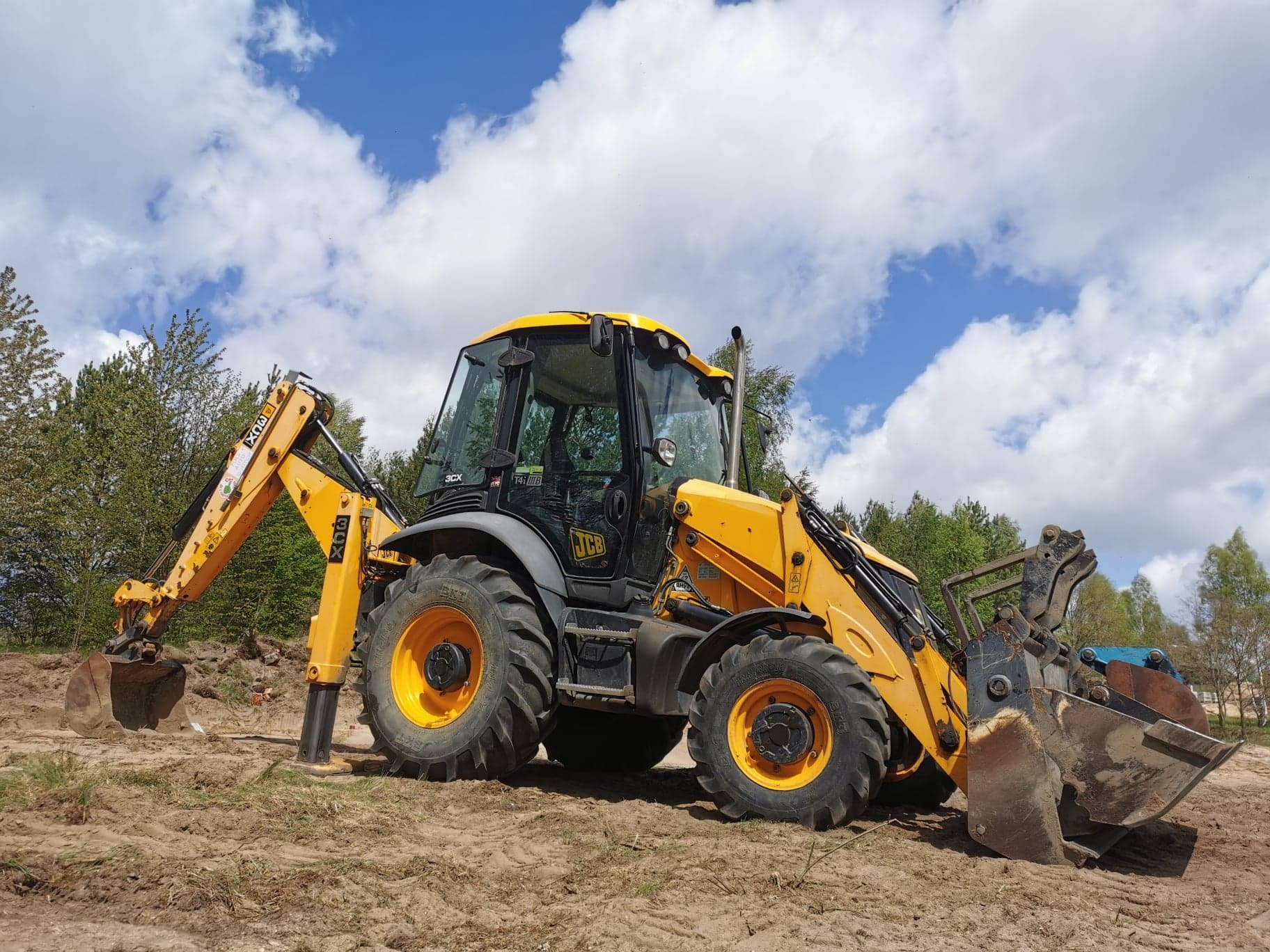 Żółto-szara koparko-ładowarka JCB na piaszczystym gruncie, z widocznym łyżką z przodu i ramieniem kopiącym z tyłu, na tle błękitnego nieba z chmurami i drzew