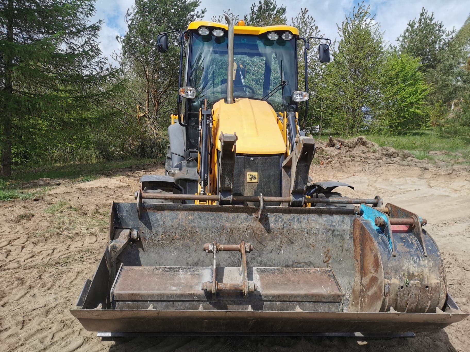 Żółta ładowarka JCB z przodu na piaszczystym terenie z drzewami w tle, widoczne ślady opon na ziemi.