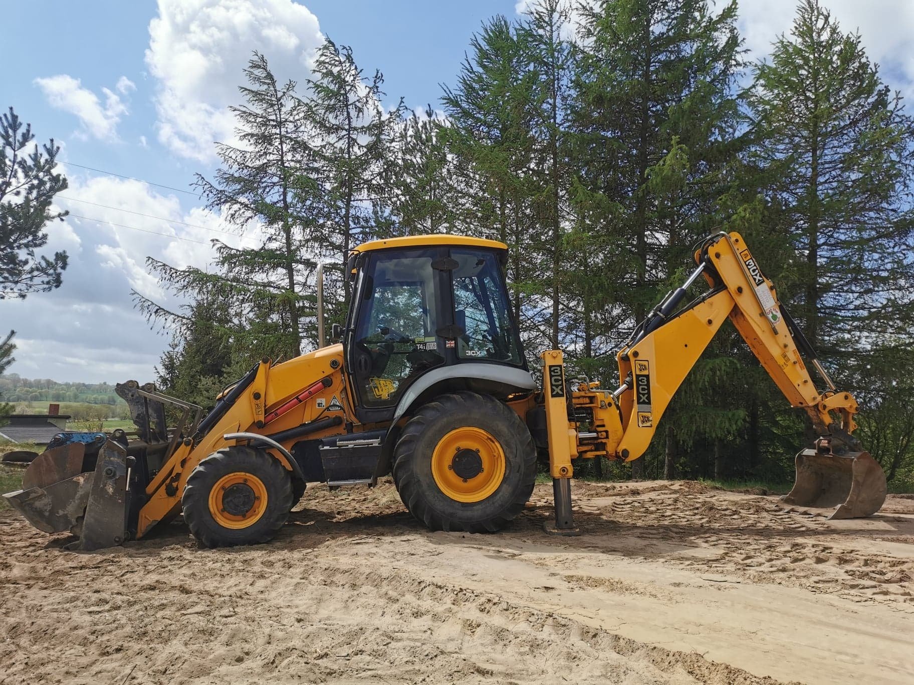 Żółta koparko-ładowarka JCB 3CX na piaszczystym gruncie, w tle wysokie sosny i błękitne niebo z białymi chmurami.