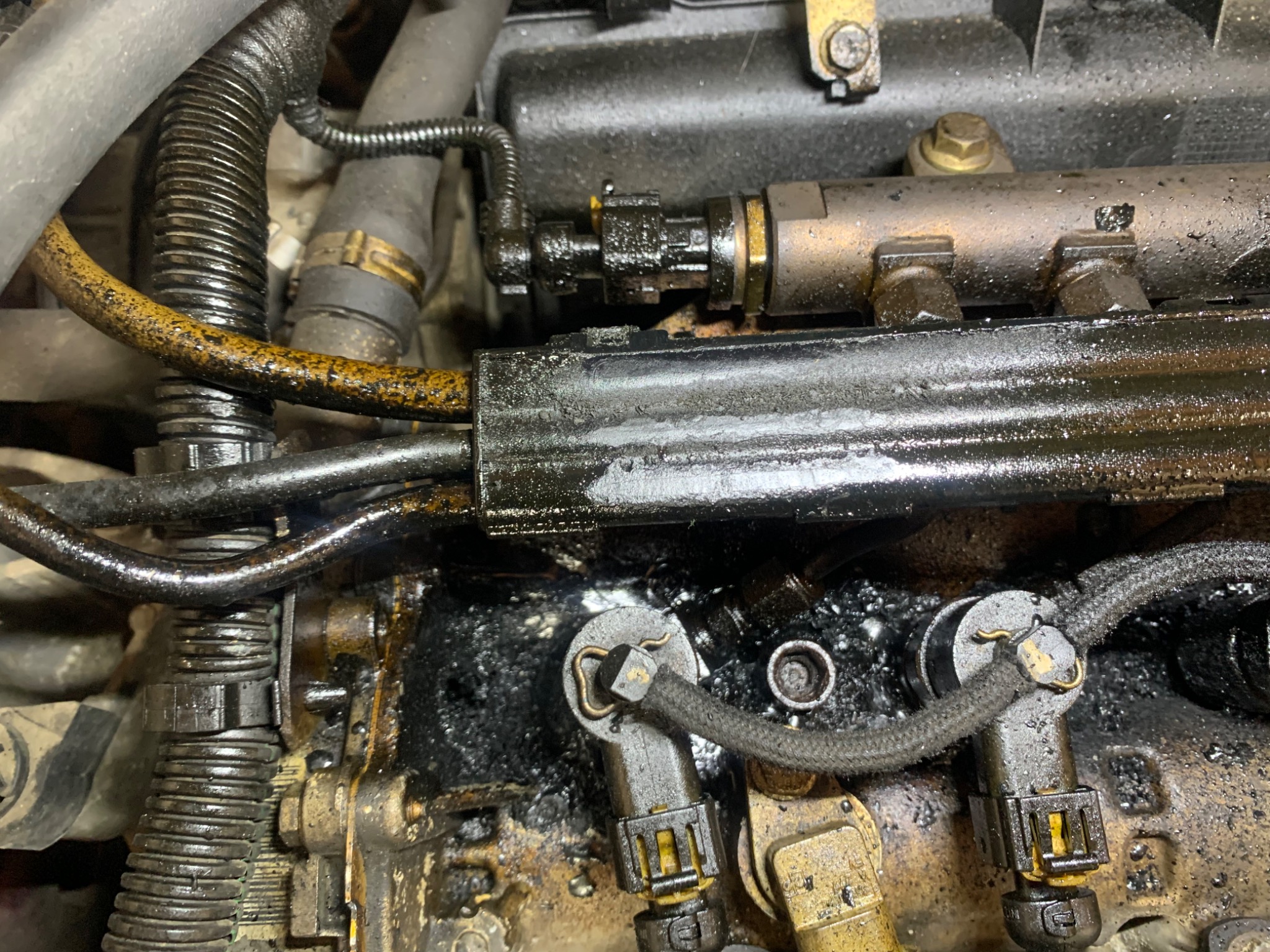 Zbliżenie na zabrudzony silnik samochodowy z widocznymi przewodami, wtryskiwaczami i elementami metalowymi, pokryty warstwą oleju i brudu.