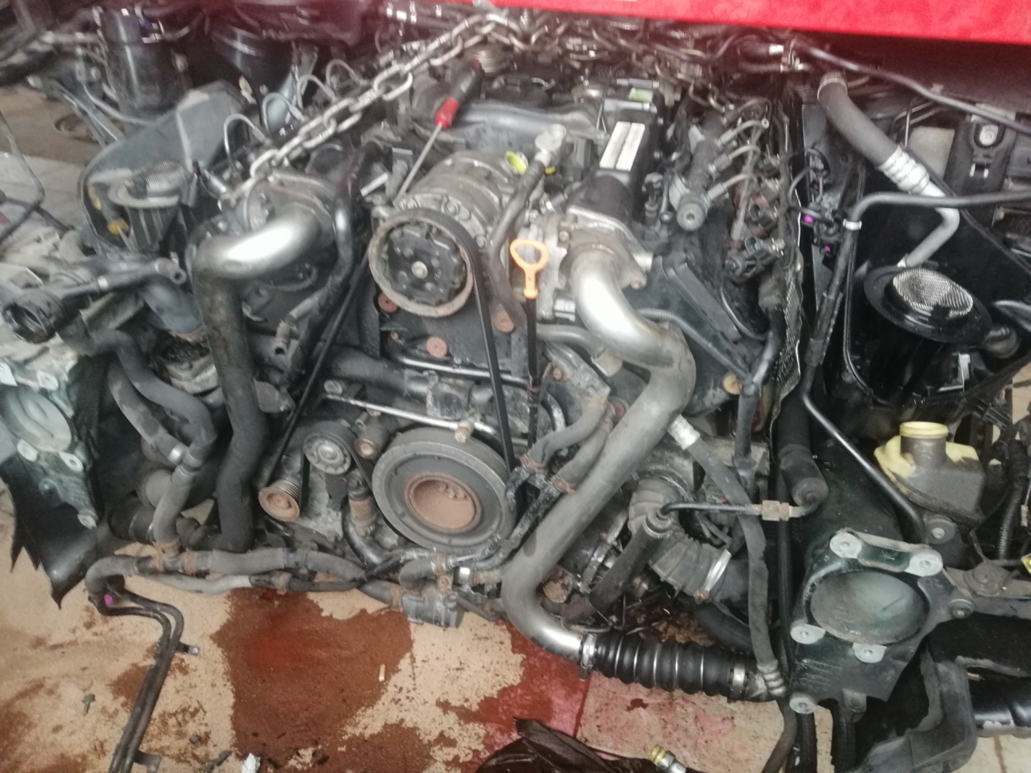 Widok na silnik samochodowy podczas naprawy, z łańcuchem podtrzymującym i widocznymi śladami wycieków płynów eksploatacyjnych na podłożu.