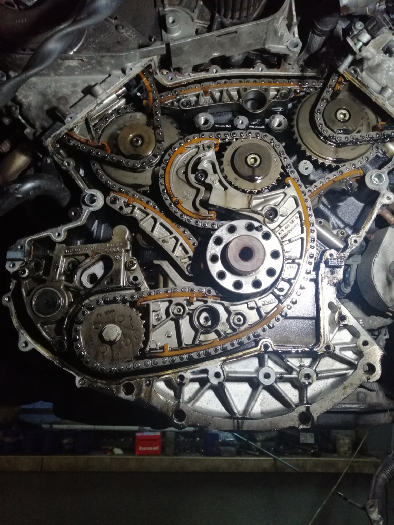 Otwarty silnik samochodowy z widocznymi łańcuchami rozrządu, zębatkami i elementami mocującymi, widoczne ślady oleju, w tle czerwone pudełko Bosal.