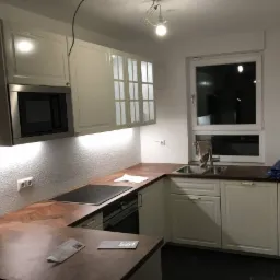 Jasna kuchnia z drewnianymi blatami w jodełkę, białymi szafkami i wbudowanym sprzętem AGD, widoczny fragment instalacji elektrycznej na suficie.