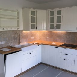 Montaż mebli Szczecin - Jasna kuchnia z białymi szafkami, drewnianym blatem i wzorzystymi płytkami na ścianie, oświetlona listwą LED pod szafkami wiszącymi.