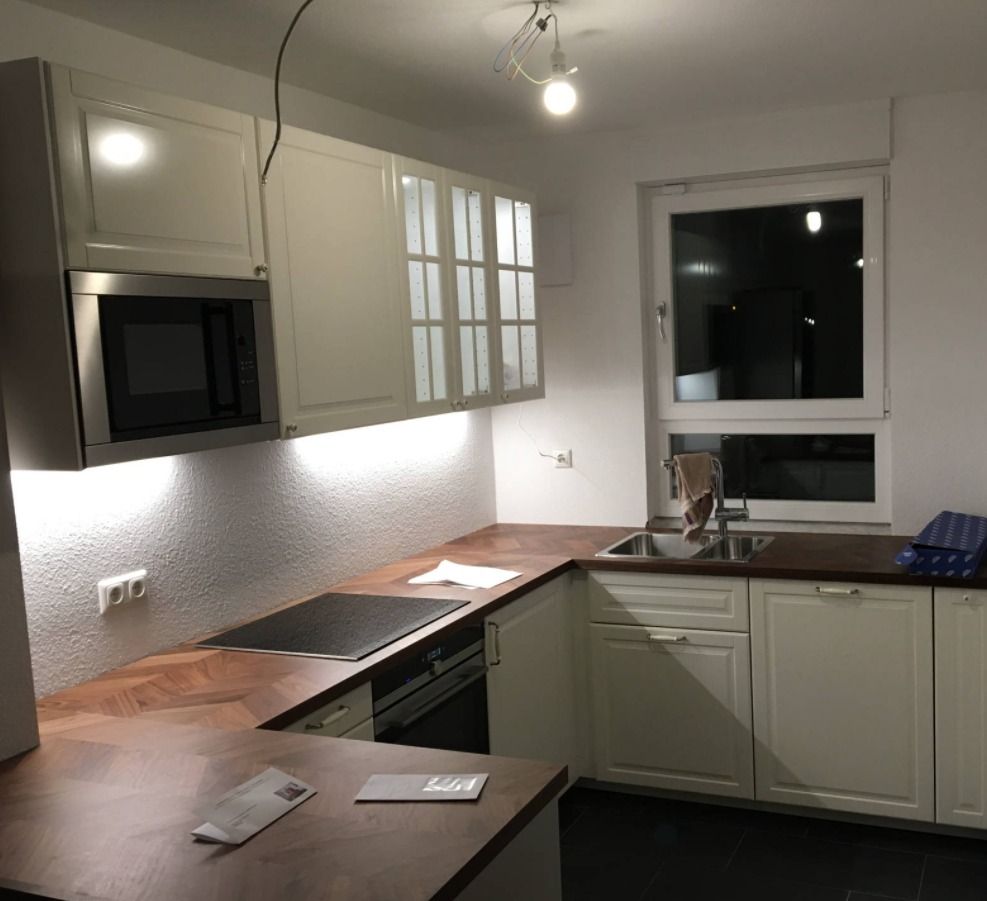 Jasna kuchnia z drewnianymi blatami w jodełkę, białymi szafkami i wbudowanym sprzętem AGD, widoczny fragment instalacji elektrycznej na suficie.