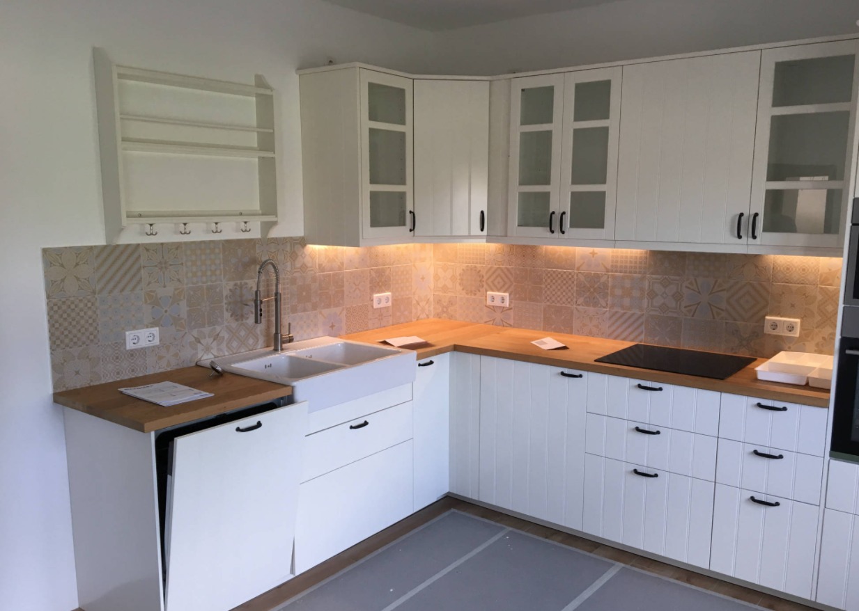 Jasna kuchnia z białymi szafkami, drewnianym blatem i wzorzystymi płytkami na ścianie, oświetlona listwą LED pod szafkami wiszącymi.