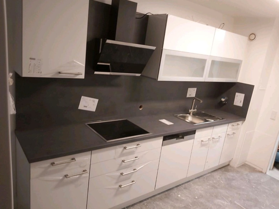 Nowoczesna kuchnia z białymi szafkami i szarym blatem, czarny okap nad płytą indukcyjną, widoczne naklejki na szafkach.