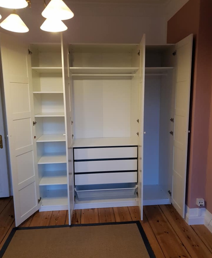 Biała trzydrzwiowa szafa z otwartymi drzwiami, widoczne półki, drążek na ubrania i trzy szuflady z czarnymi uchwytami, na podłodze drewniany parkiet i dywan.