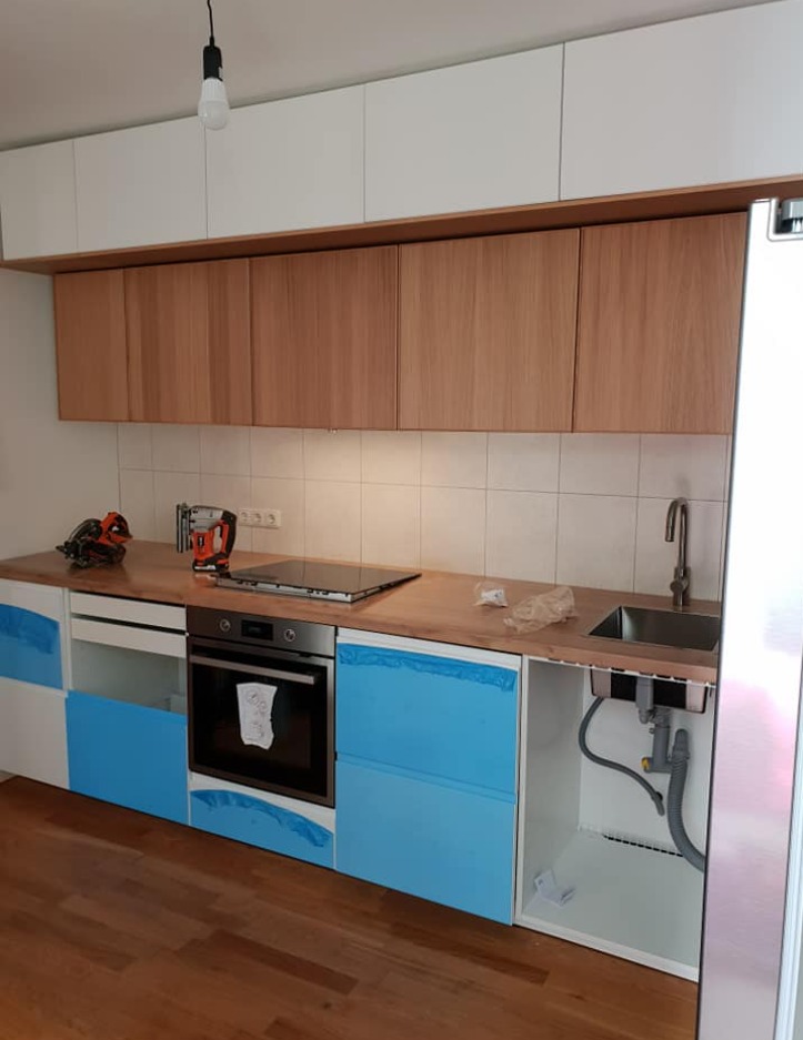 Kuchnia w trakcie montażu: drewniany blat, białe i niebieskie fronty szafek zabezpieczone folią, widoczne narzędzia i podłączenie zlewu.