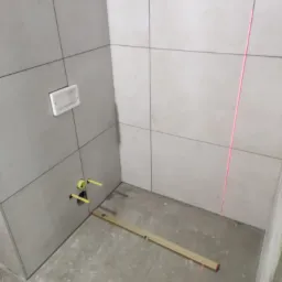 Szare płytki ścienne w trakcie układania w rogu łazienki, widoczny poziomica i laser krzyżowy wyznaczający linię.