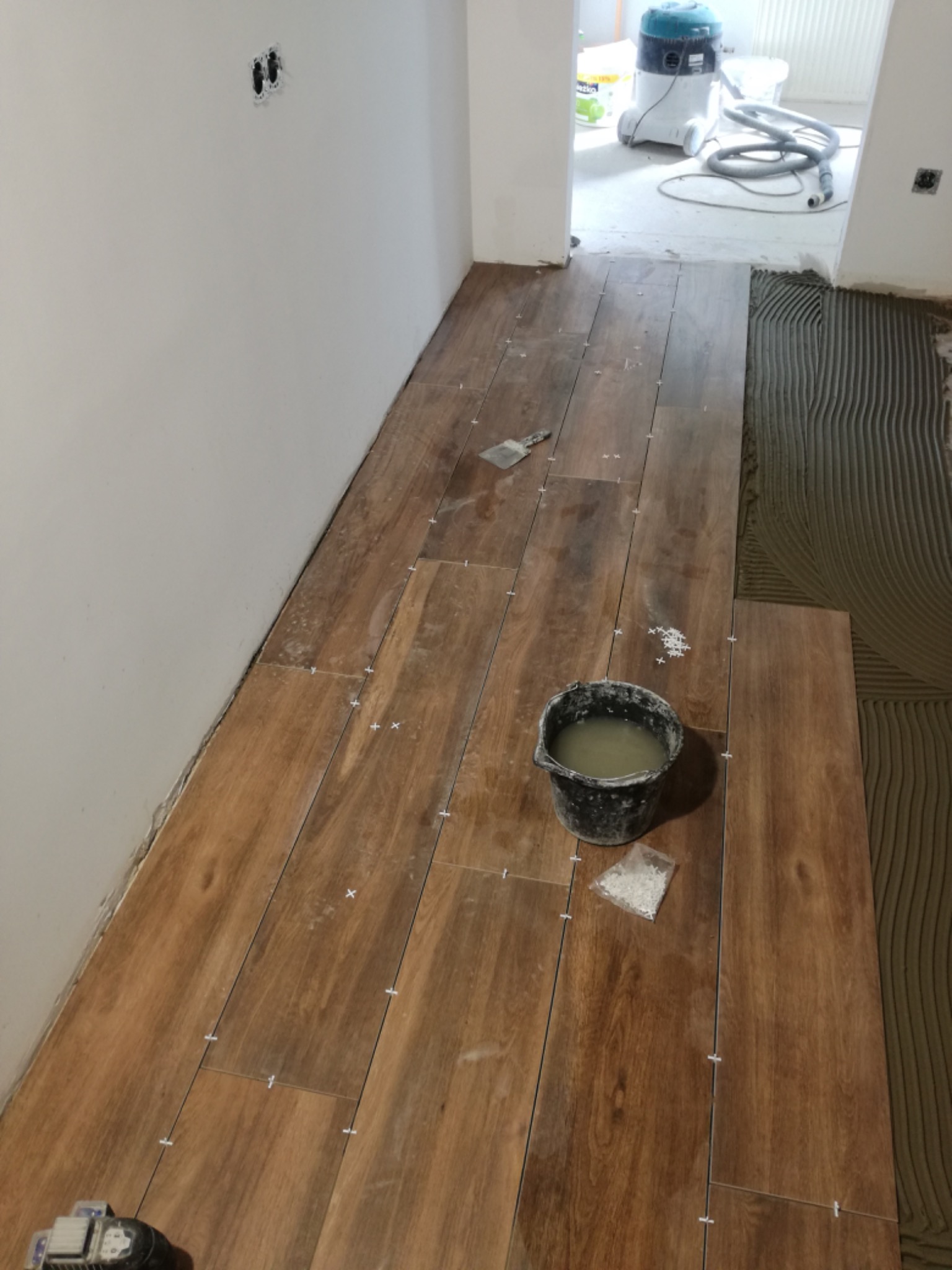 Układanie podłogi z płytek imitujących drewno, widoczne krzyżyki dystansowe, wiadro z wodą i paczka z krzyżykami, w tle odkurzacz przemysłowy.