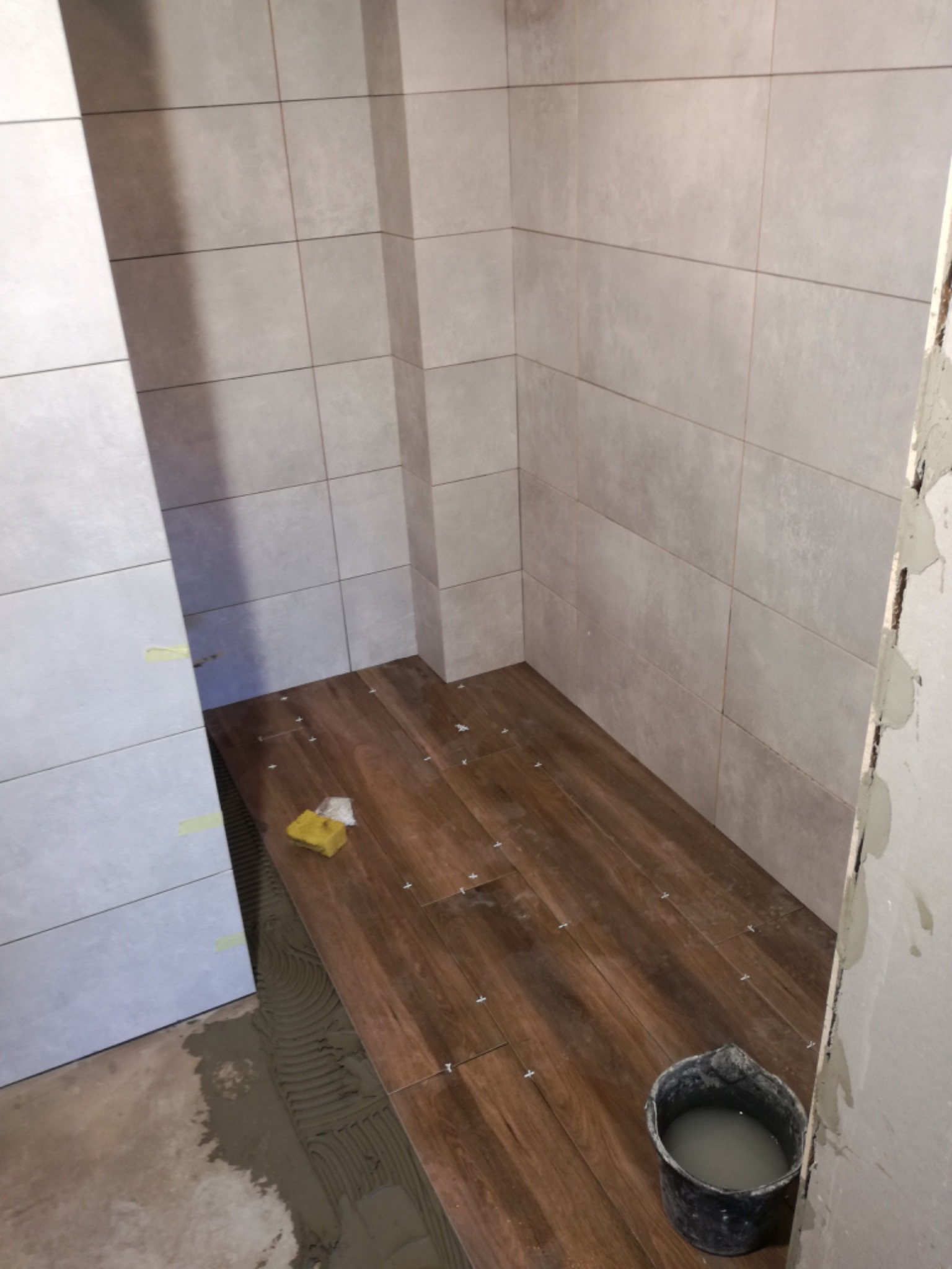 Wnętrze łazienki w trakcie układania brązowych płytek podłogowych imitujących drewno, szare płytki na ścianach, widoczne krzyżyki dystansowe i wiadro z zaprawą.