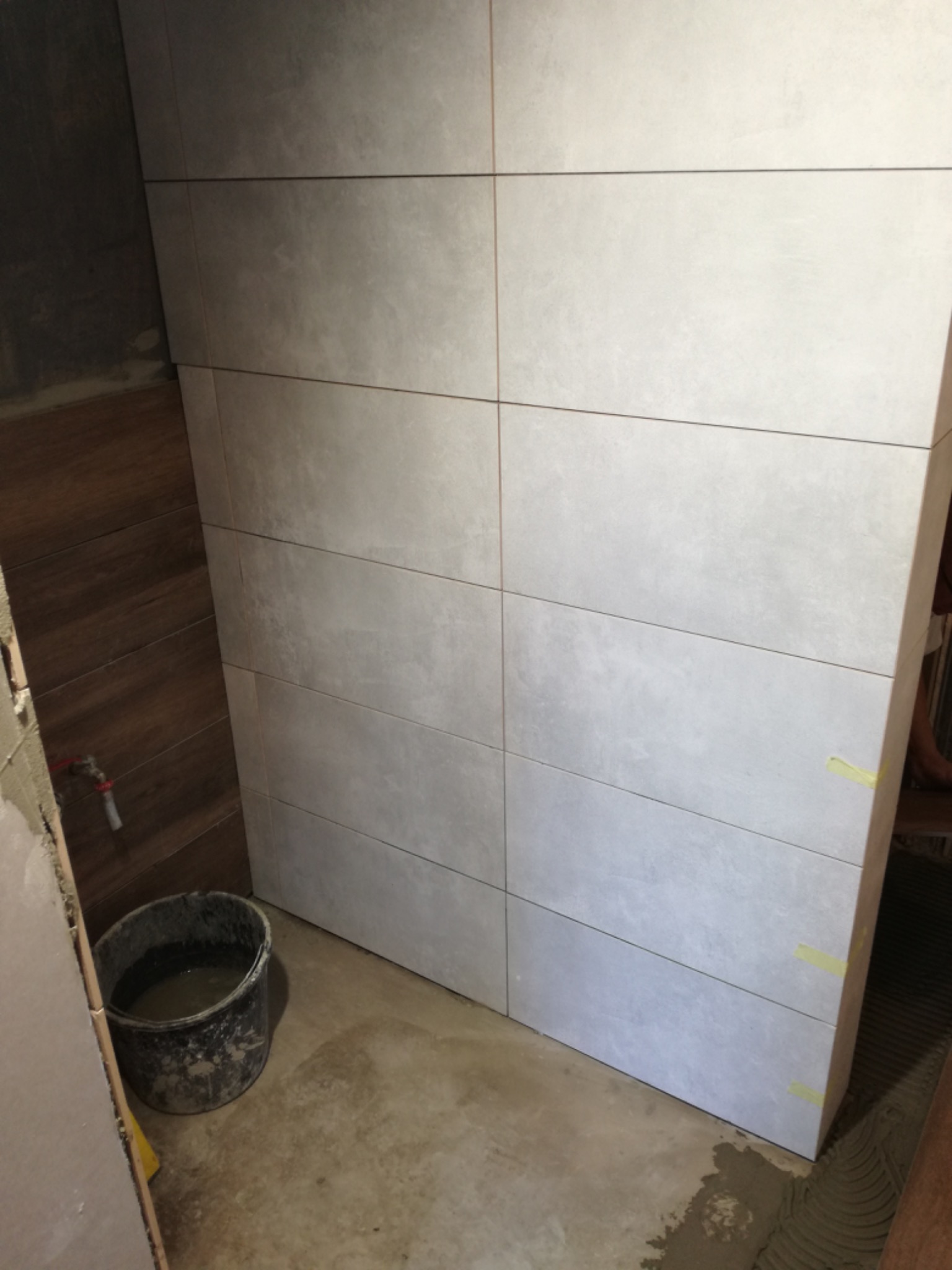 Ściana w trakcie układania płytek w łazience, widoczne szare, prostokątne płytki, wiadro z zaprawą i fragment drewnianej ściany obok.