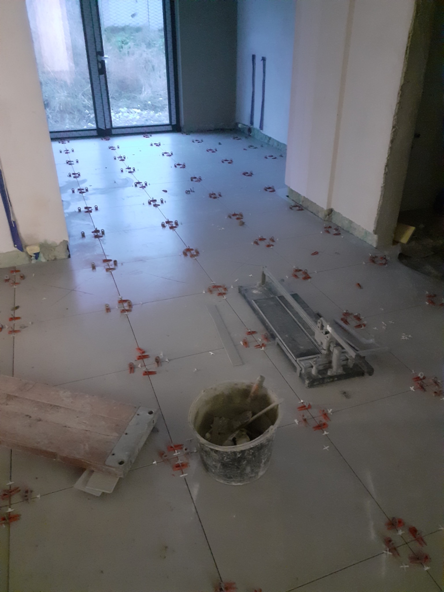 Szare płytki podłogowe w trakcie układania, widoczne krzyżyki dystansowe, wiadro z zaprawą i przecinarka do płytek.