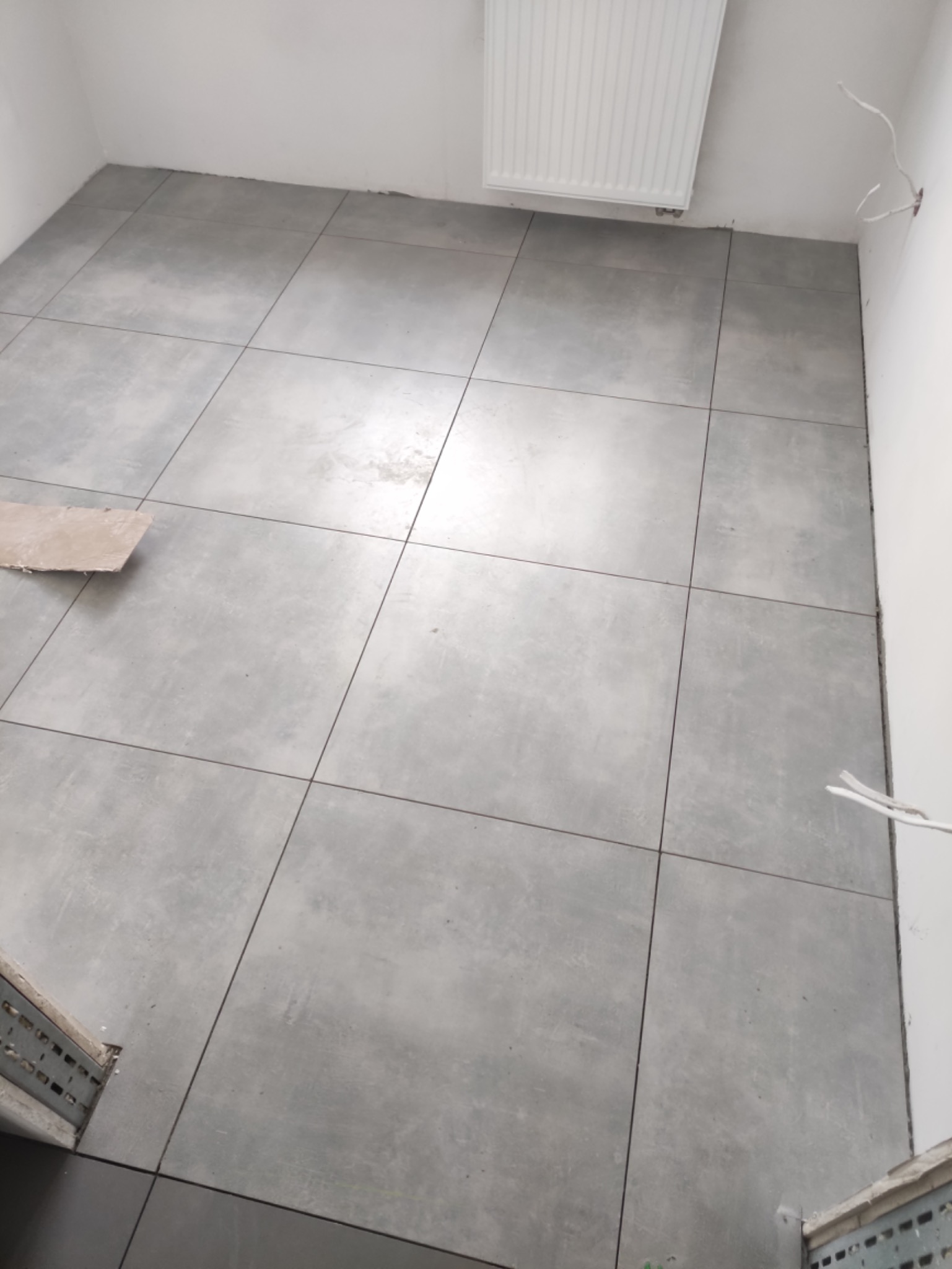 Ułożone szare płytki podłogowe w trakcie prac wykończeniowych, widoczny grzejnik i wystające przewody elektryczne.