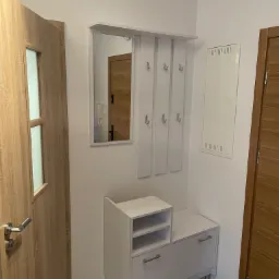 Nowoczesny przedpok&oacute;j z białymi meblami: szafka na buty z siedziskiem, panel z lustrem i wieszakami, drewniane drzwi z przeszkleniem, białe ściany, podłoga imitująca drewno.