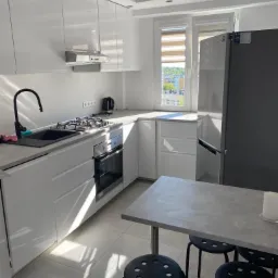 Jasna, nowoczesna kuchnia z białymi, błyszczącymi szafkami, czarną armaturą i kuchenką gazową, szarym blatem i stołem z trzema czarnymi stołkami, widok na okno z roletami.