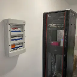 Rozdzielnica elektryczna natynkowa z bezpiecznikami i wyłącznikami, obok szafa rack z patch panelem i switchami, kable poprowadzone wewnątrz szafy.