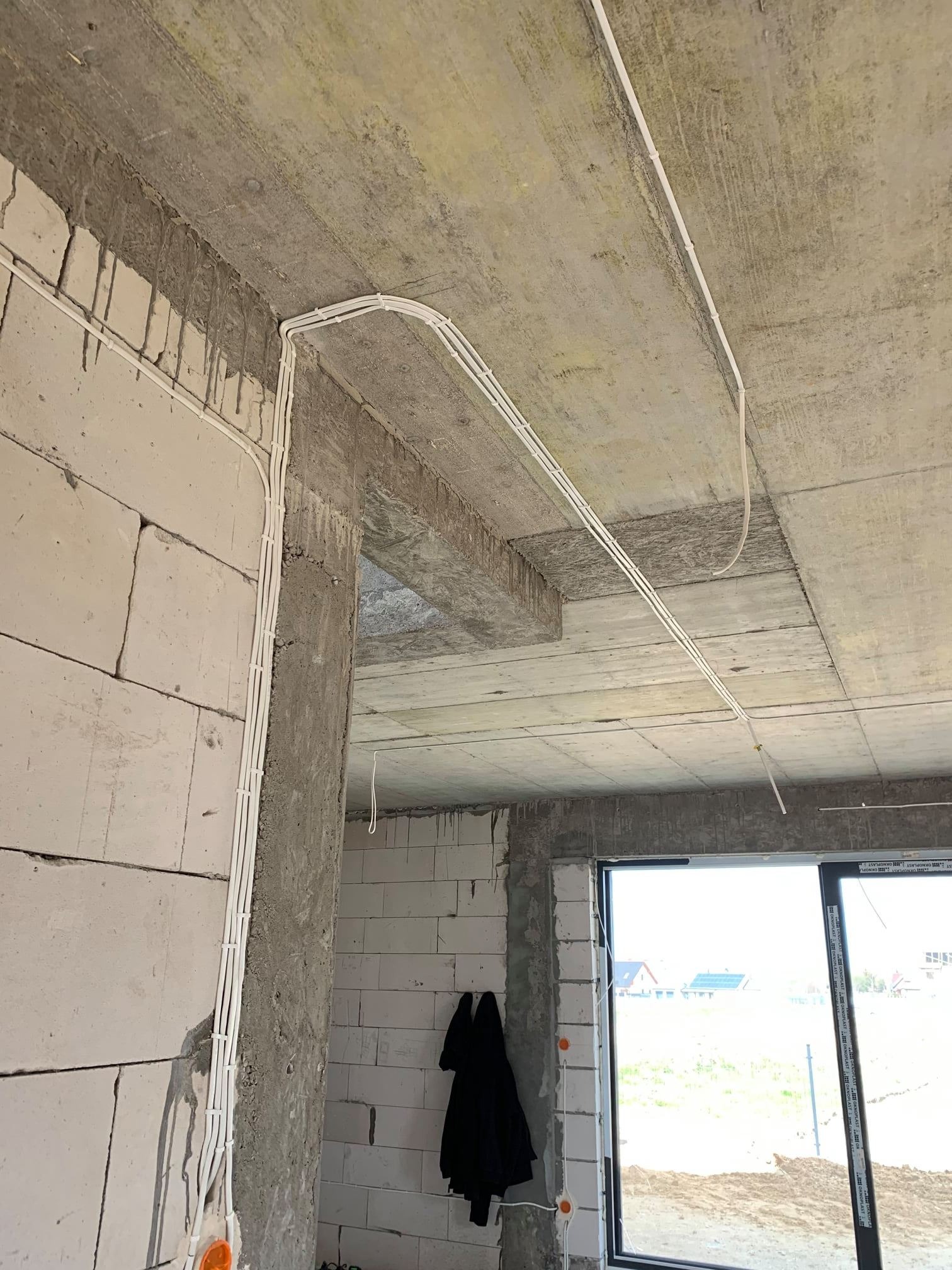 Surowy stan budynku z widoczną instalacją elektryczną w białych rurkach przytwierdzonych do ścian i sufitu z betonu komórkowego, okno z widokiem na zewnątrz.