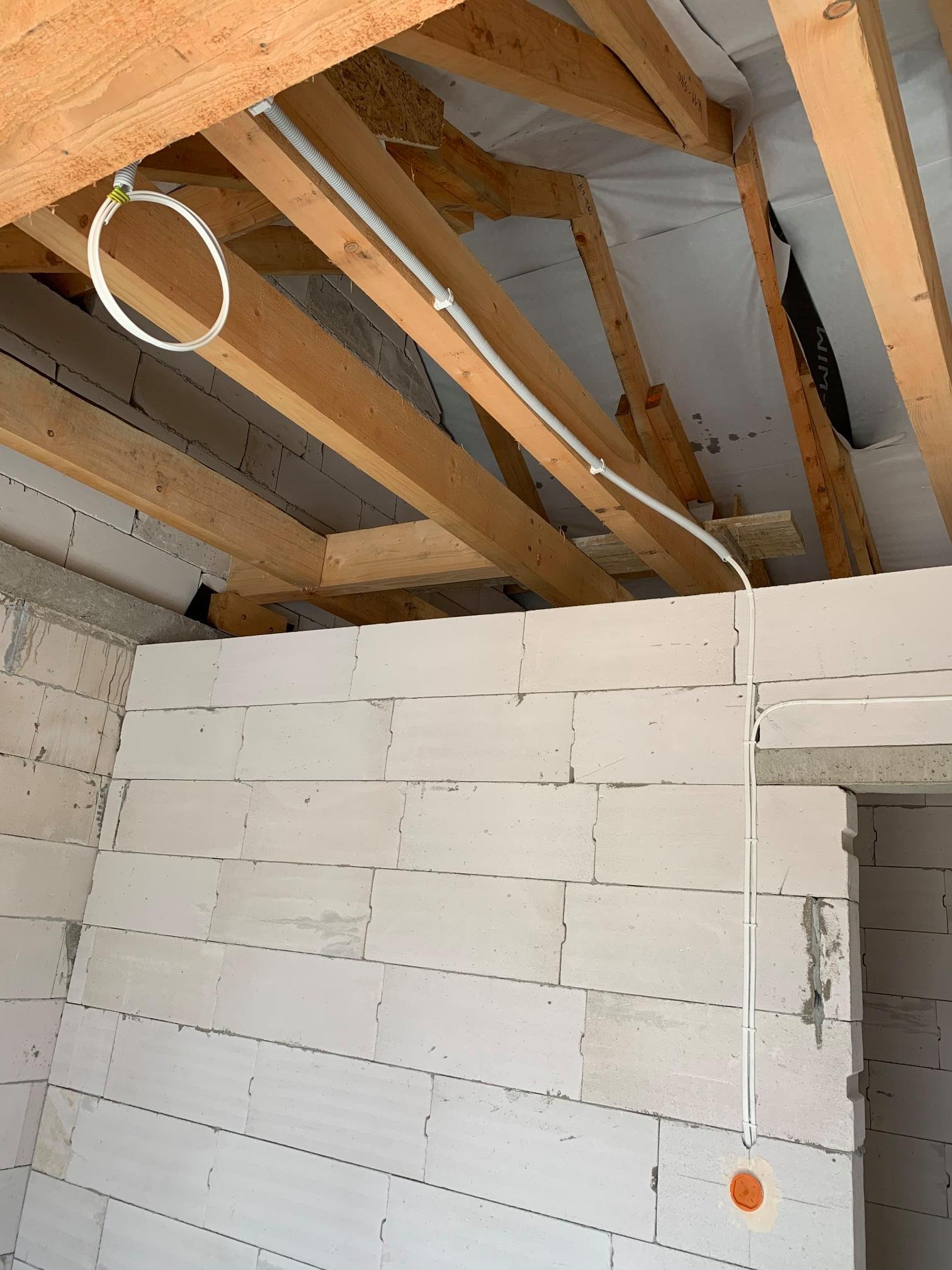 Wewnętrzna instalacja elektryczna w surowym stanie: białe rury prowadzone po drewnianej konstrukcji dachu i ścianach z białych bloczków, widoczne puszki instalacyjne.