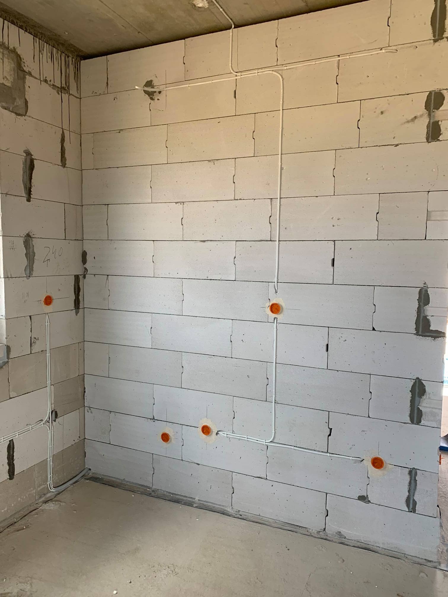 Surowy mur z betonu komórkowego z widoczną instalacją elektryczną w rurkach PCV, pomarańczowe puszki pod gniazdka, ślady po zaprawie w szczelinach, napis '240' na ścianie.