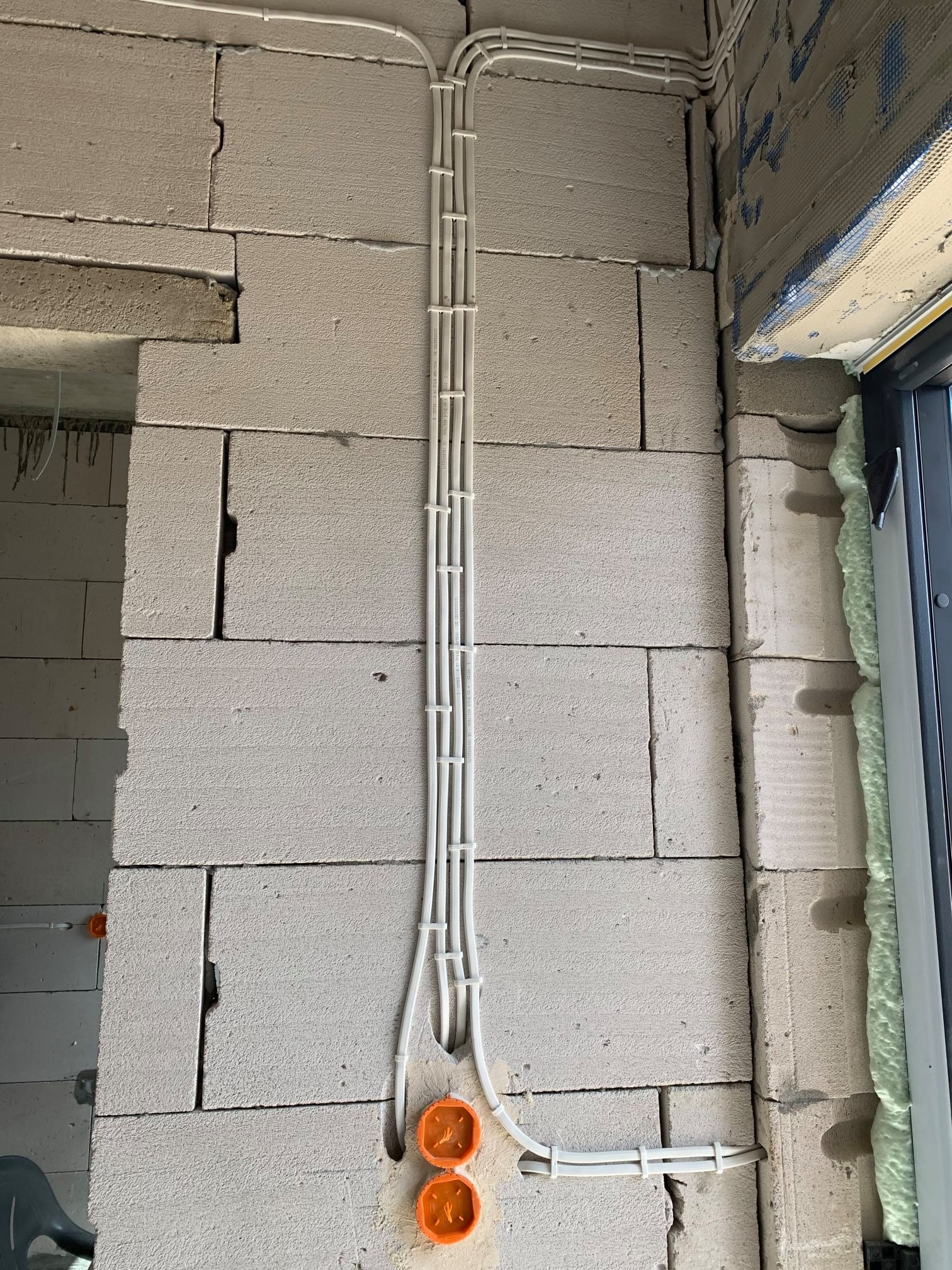 Białe przewody elektryczne ułożone pionowo w korytkach na ścianie z białych bloczków betonowych, widoczne pomarańczowe puszki instalacyjne i fragment okna z pianką montażową.