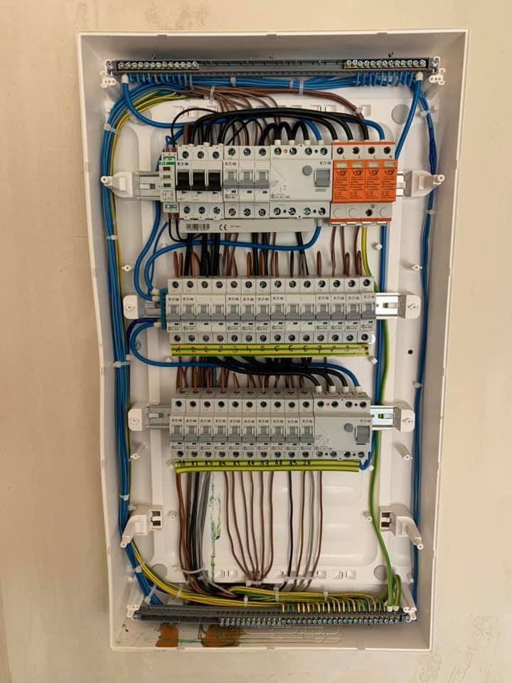 Otwarta skrzynka rozdzielcza z widocznymi bezpiecznikami, przewodami w różnych kolorach i listwami zaciskowymi, zamontowana na jasnej ścianie.