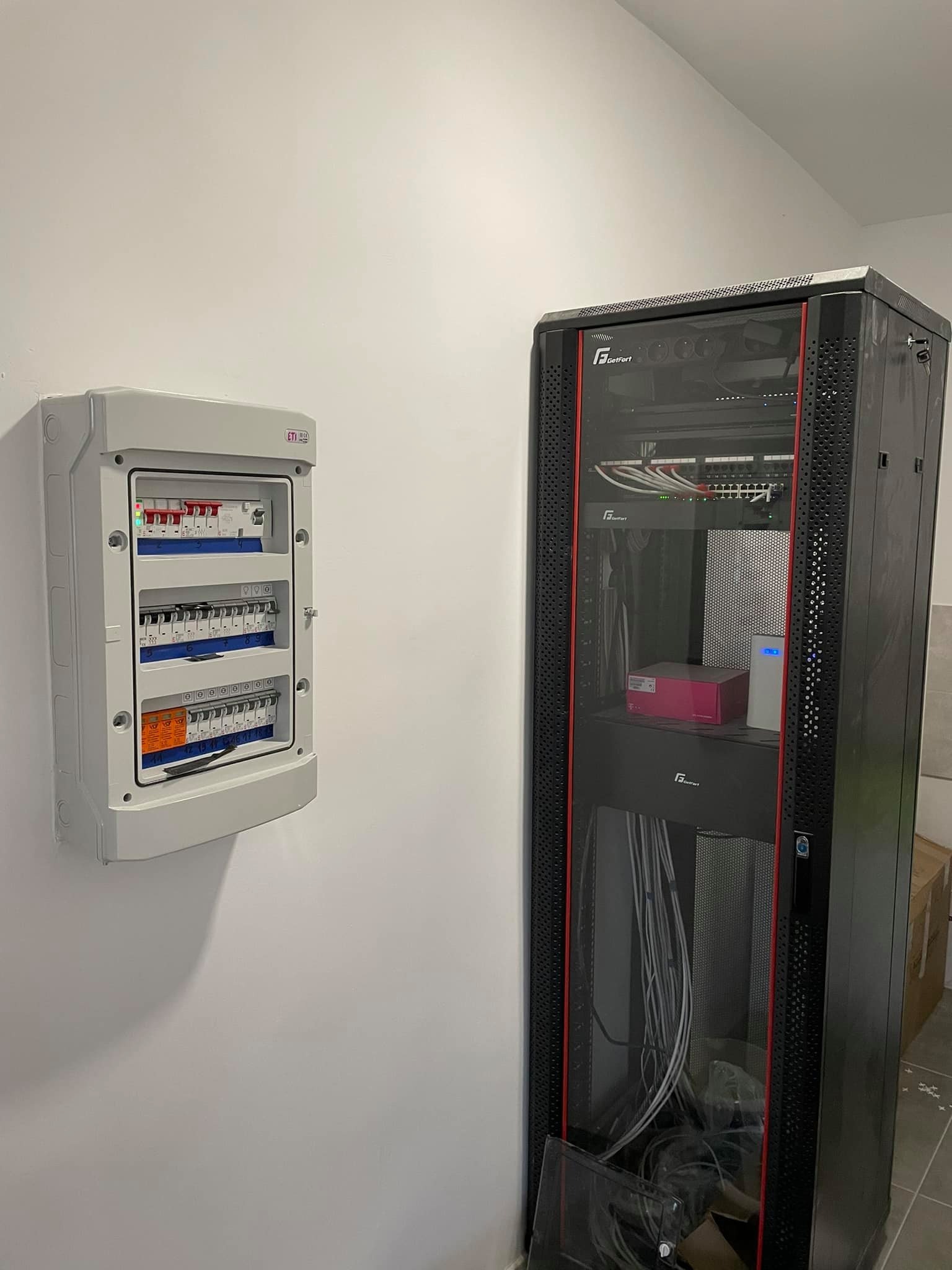 Rozdzielnica elektryczna natynkowa z bezpiecznikami i wyłącznikami, obok szafa rack z patch panelem i switchami, kable poprowadzone wewnątrz szafy.