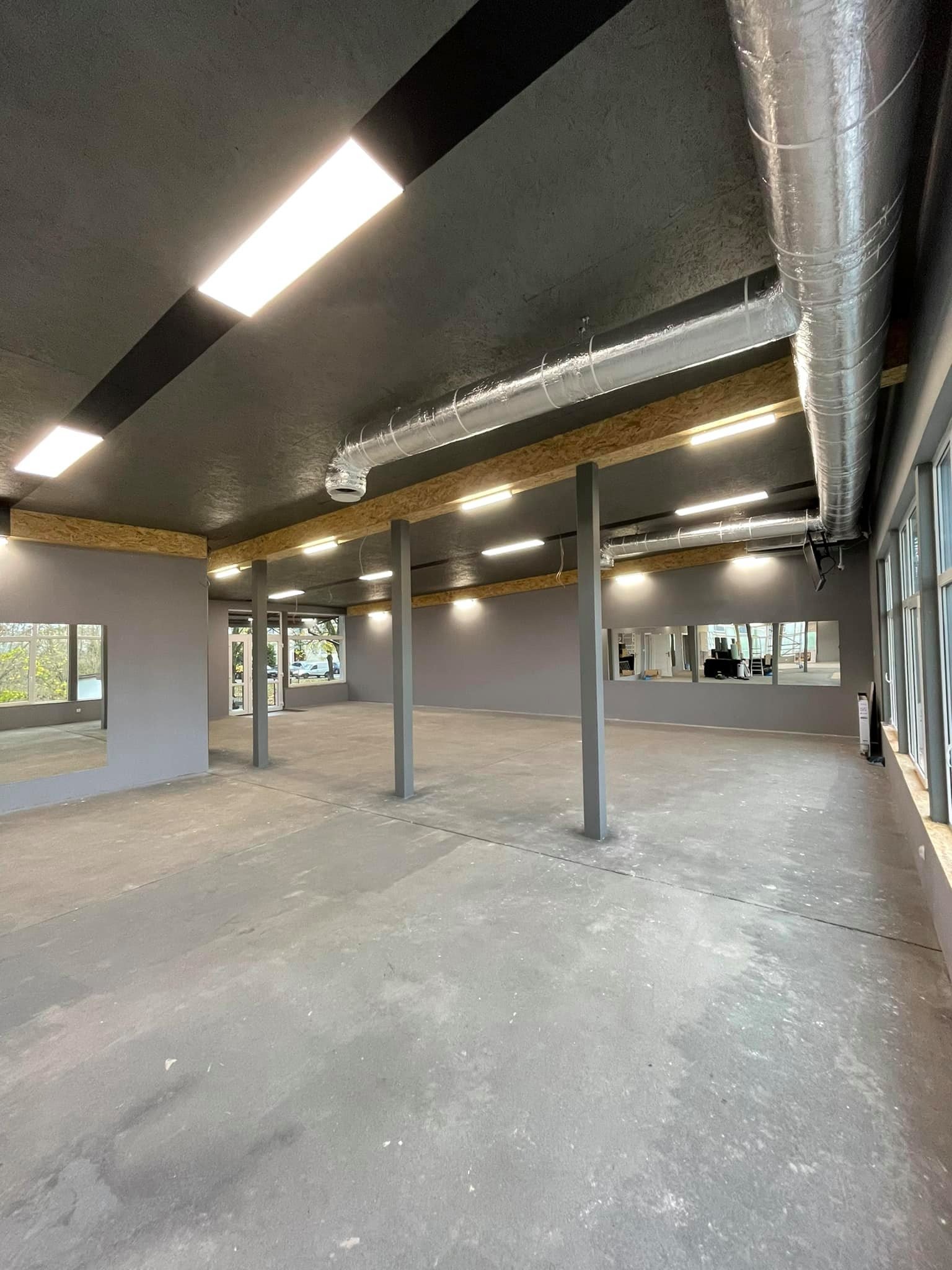 Wnętrze pustego lokalu z szarą podłogą i ścianami, widoczne instalacje wentylacyjne pod sufitem, oświetlenie LED i kolumny wspierające strop.