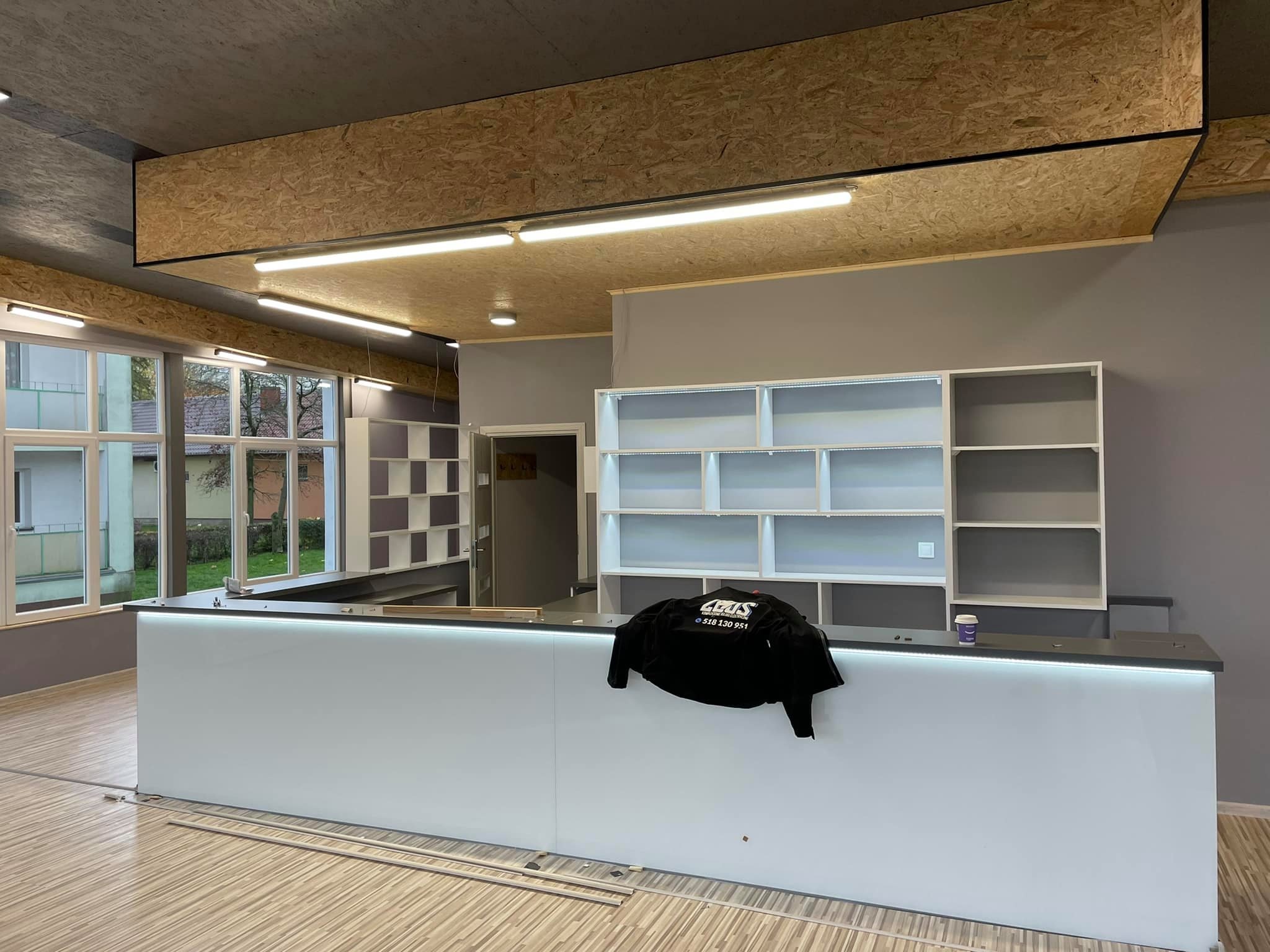 Wykończone wnętrze lokalu z białą ladą z podświetleniem LED, regały w tle i surowy sufit z płyt OSB, czarna kurtka z logo firmy leży na ladzie.