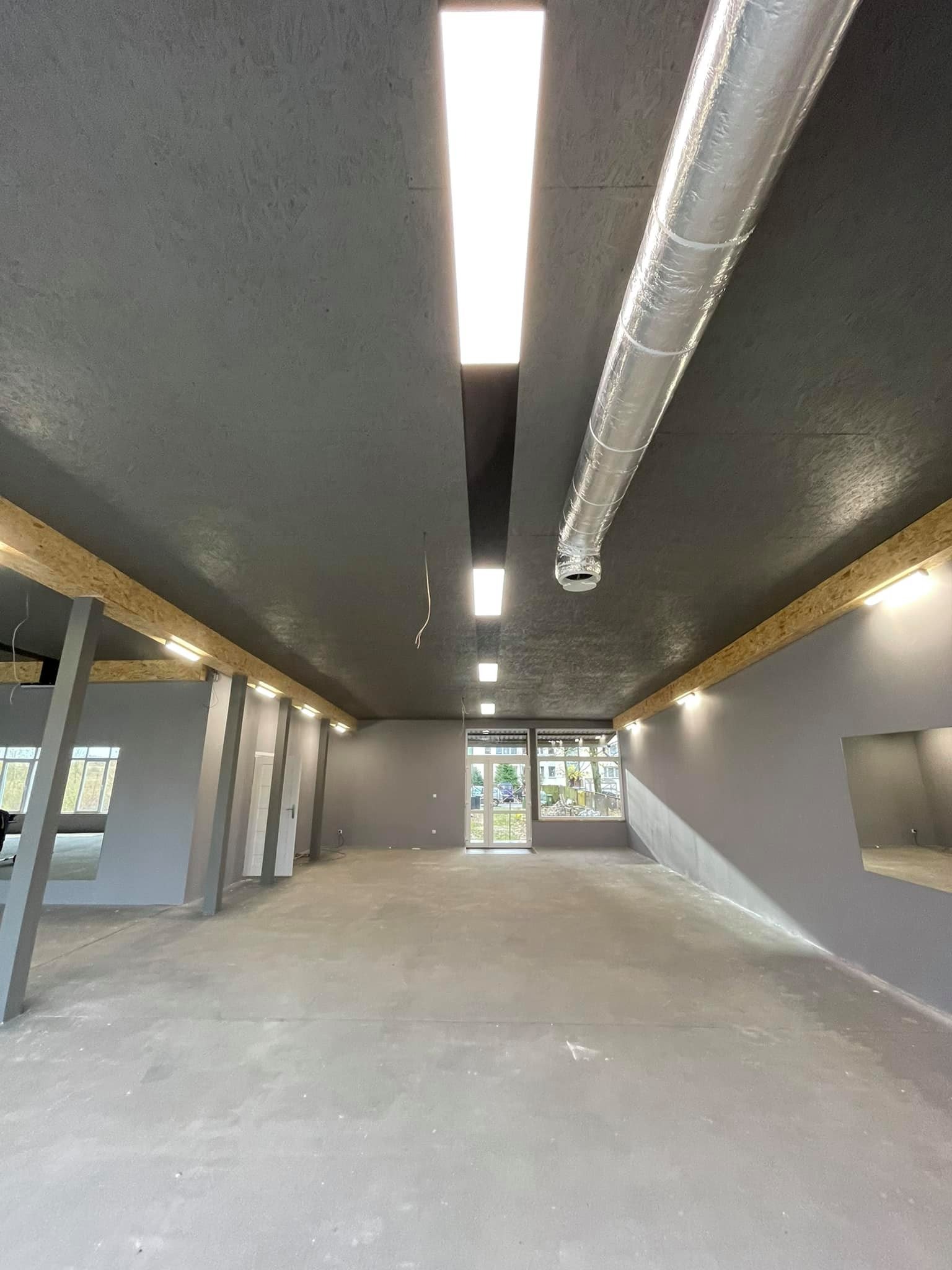 Surowe wnętrze w szarościach z widoczną instalacją wentylacyjną, drewnianymi belkami z oświetleniem i betonową podłogą, przygotowane do dalszych prac wykończeniowych.