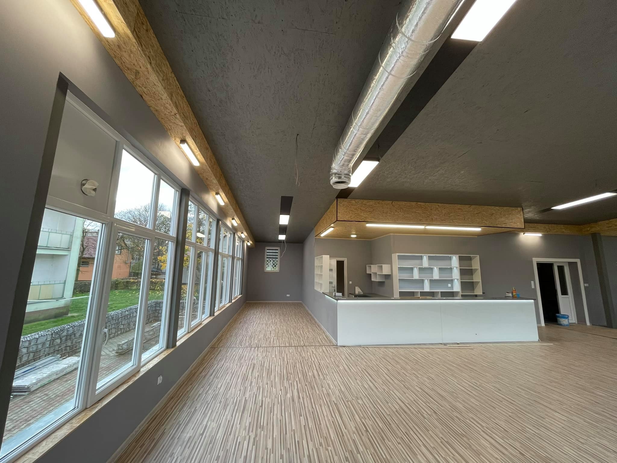 Przestronne wnętrze w szarościach z podłogą imitującą drewno, długi rząd okien z widokiem na zewnątrz, sufit z widocznymi instalacjami wentylacyjnymi i oświetleniem LED, świeżo wykończona...