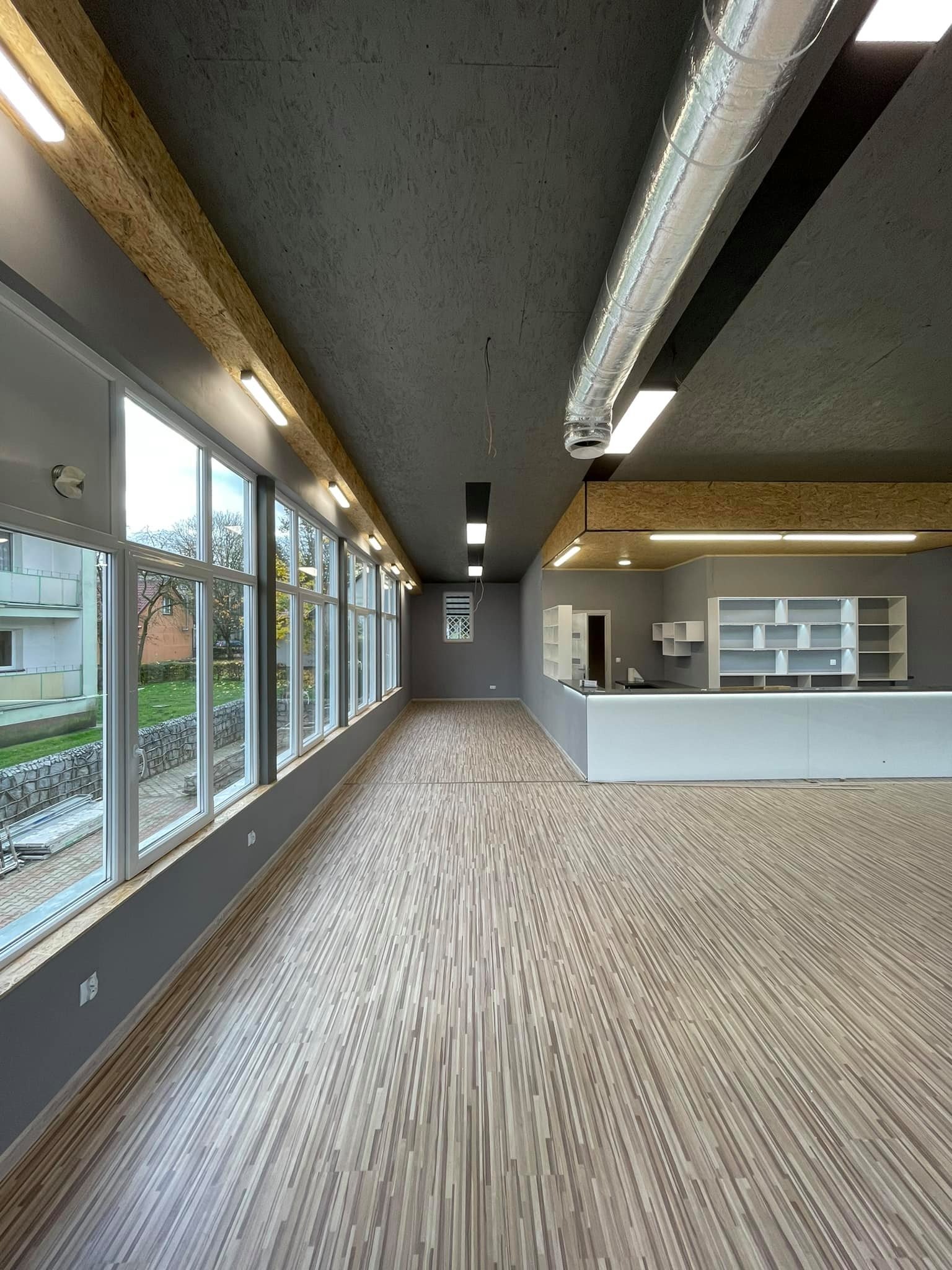 Wnętrze nowo wykończonego lokalu z drewnianą podłogą, szarymi ścianami i rzędem okien wzdłuż jednej ściany, widoczny system wentylacji pod sufitem i zabudowa barowa z półkami.