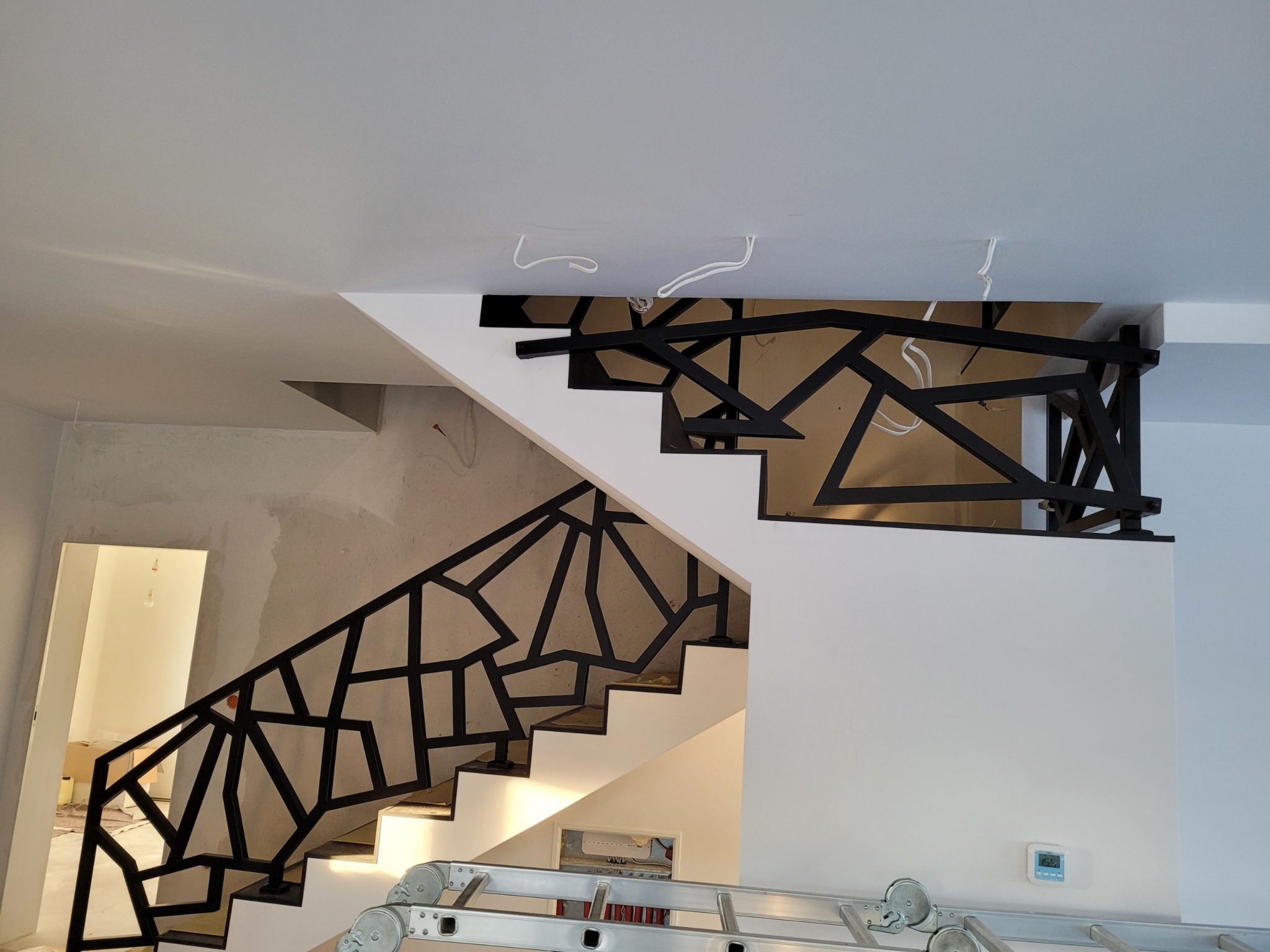 Nowoczesne schody z biało-czarną geometryczną balustradą, widok z dołu, częściowo niewykończone ściany, drabina w dolnej części kadru.