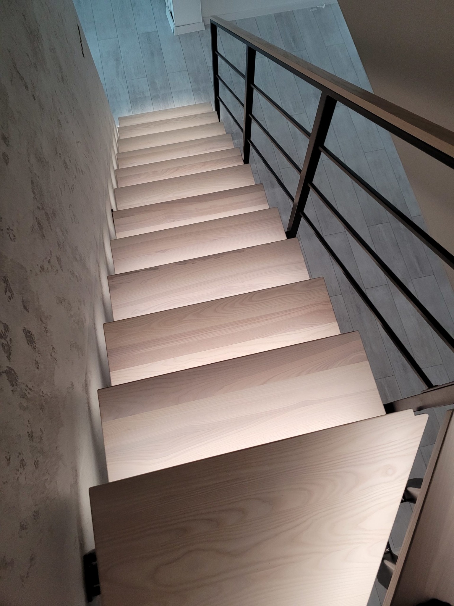 Nowoczesne schody z jasnego drewna z podświetleniem LED i czarną metalową balustradą, widok z góry.