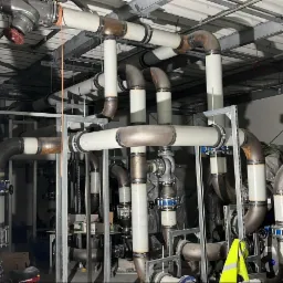 Rozbudowana instalacja rur przemysłowych z licznymi złączkami, zaworami i spawami, zamontowana pod sufitem hali.